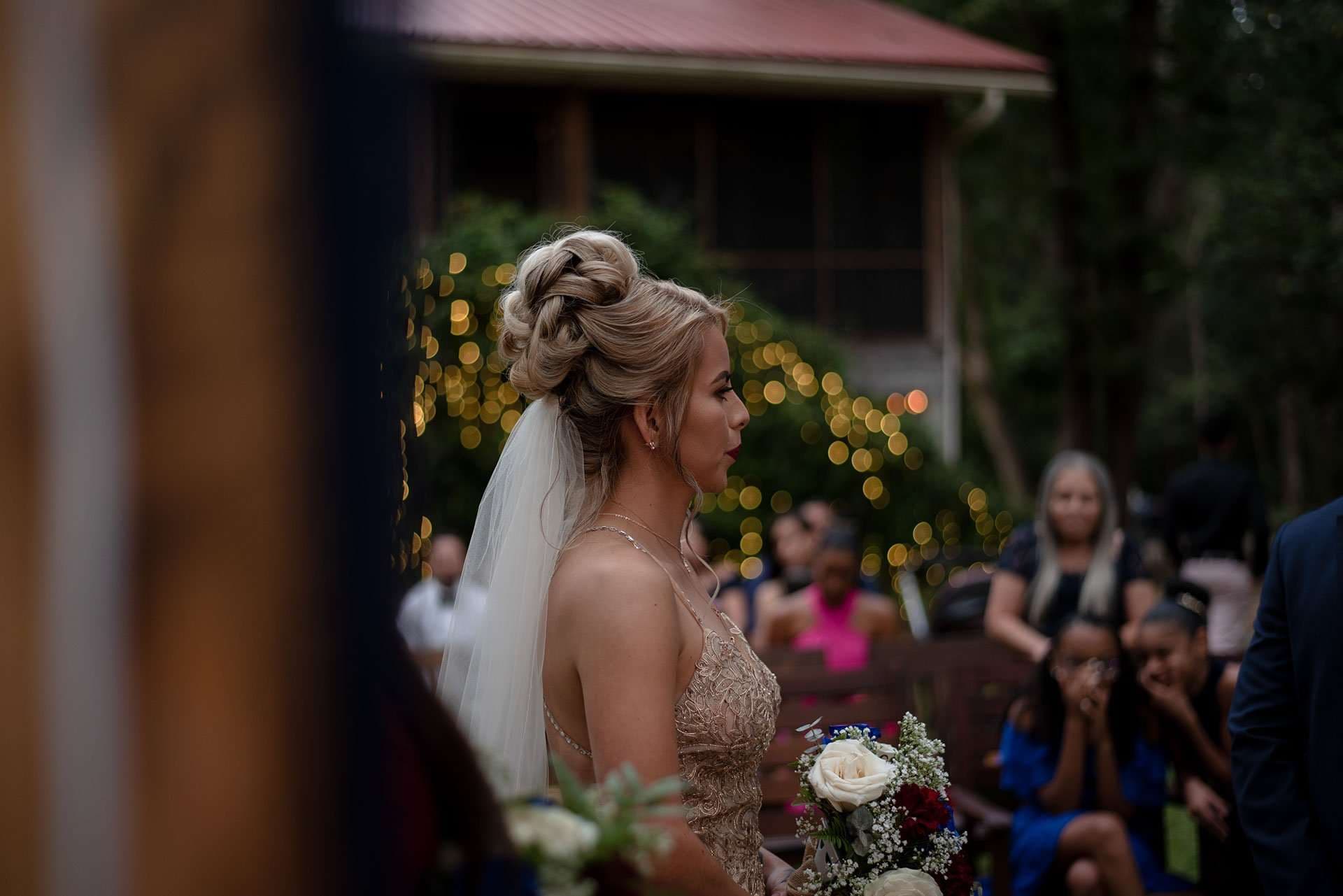 Bride in profile with an updo, veil and beaded gold gown holding a bouquet as guests watch behind her