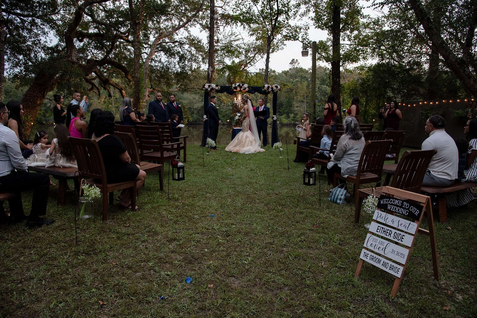 Outdoor wedding ceremony under oak trees as guests seated on wooden chairs watch a couple at a floral arch