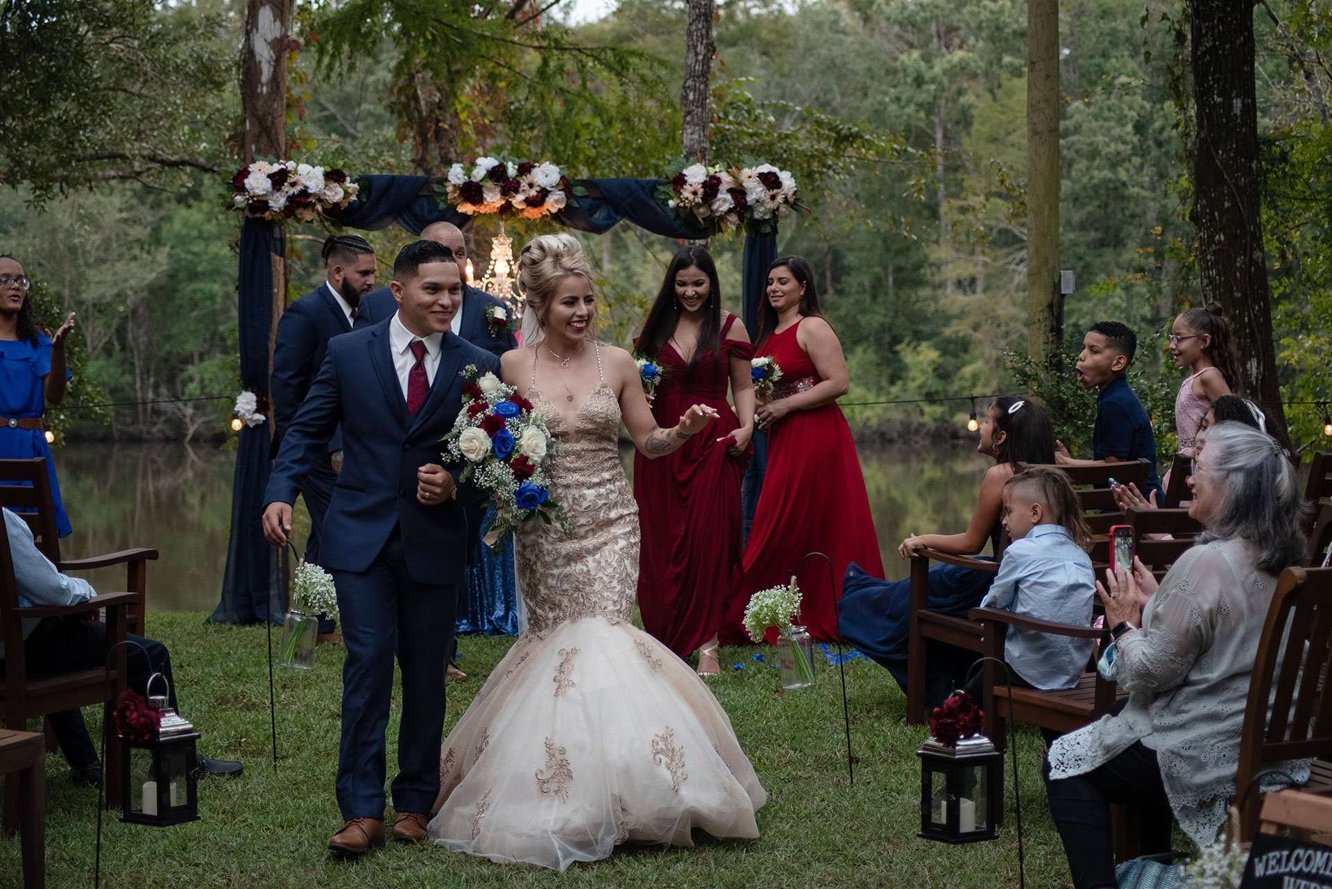 Couple walking down the aisle outdoors past seated guests, bride in a gold lace gown holding a red, white and blue bouquet