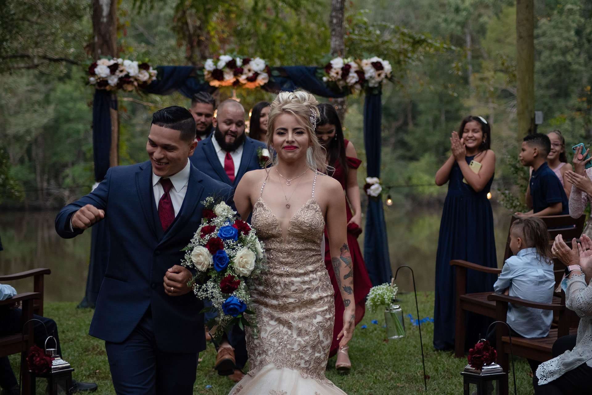 Smiling couple walking up the aisle past applauding guests, bride in a sparkling gold gown holding a blue and white bouquet