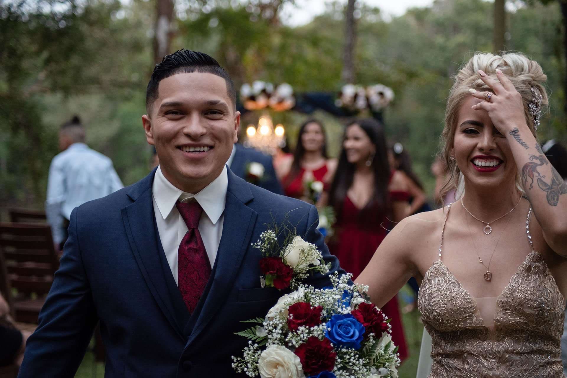 Smiling groom in navy suit holding a red and blue bouquet beside a laughing bride wiping her forehead outdoors