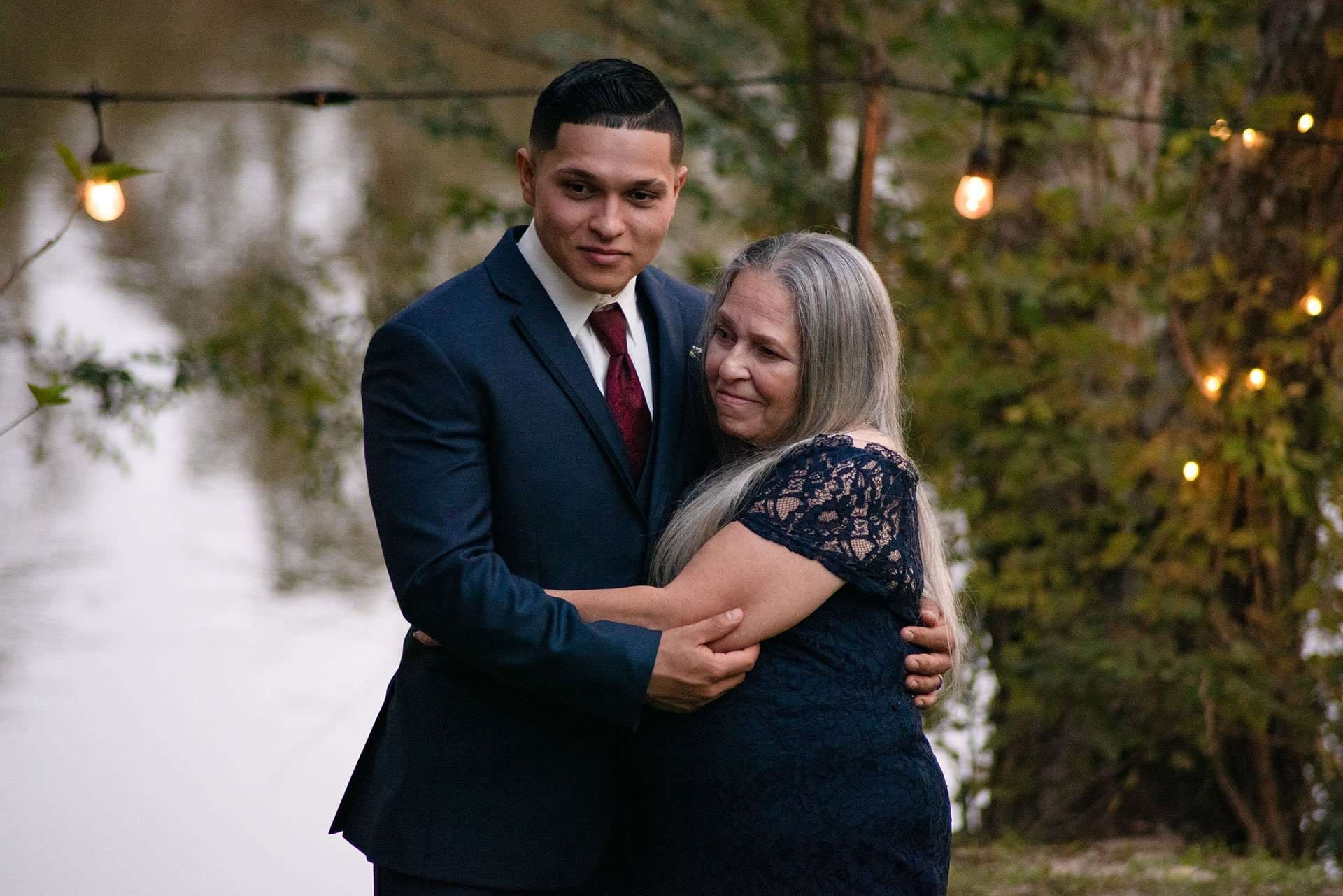 Young man in a navy suit and burgundy tie hugging an older woman in a navy lace dress beneath string lights