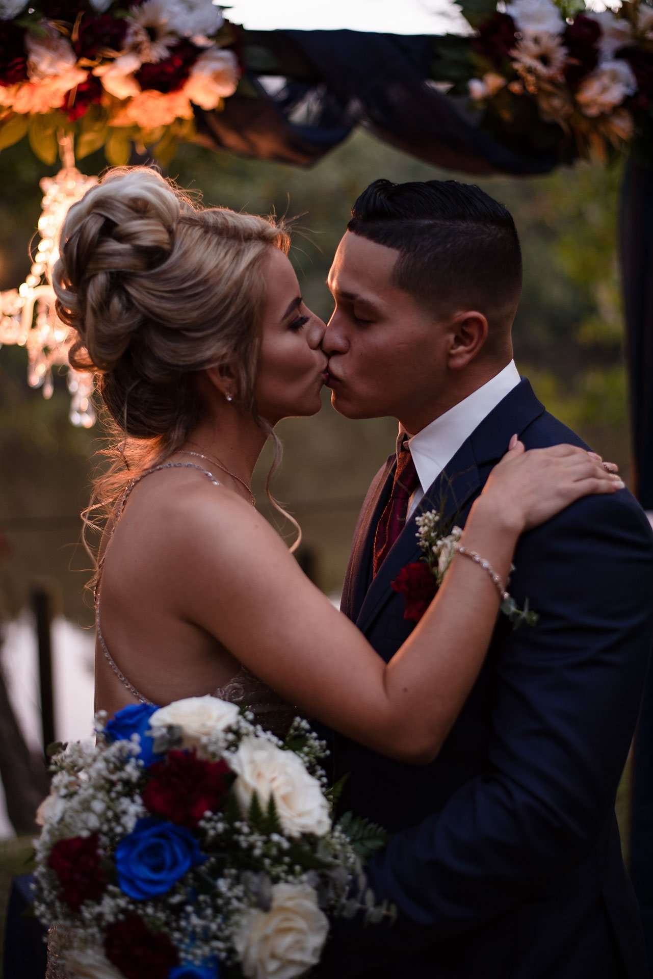 Couple kissing under a floral wedding arch at dusk, she in an updo holding a bouquet, he in a navy suit