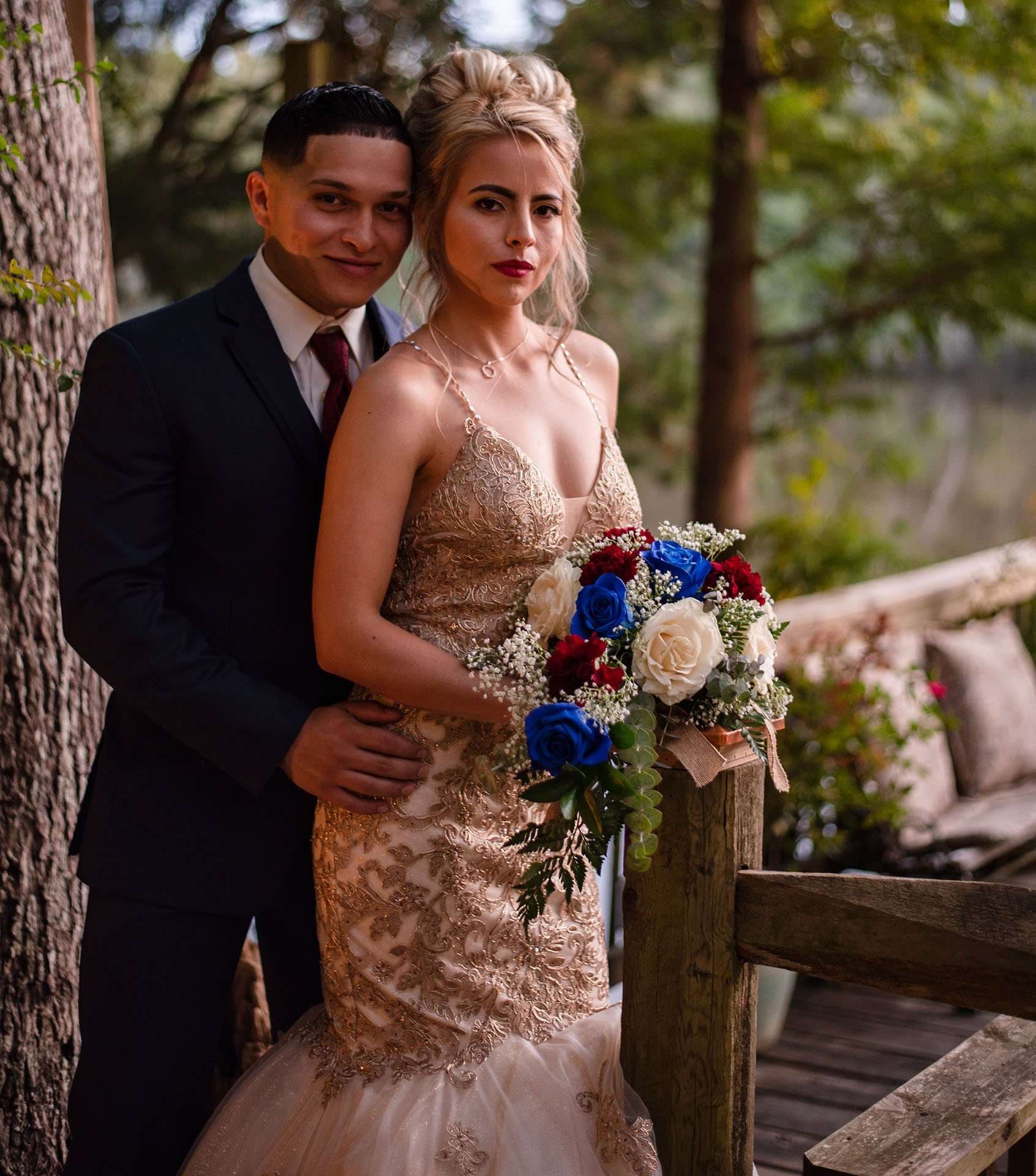 Groom in a dark suit embracing his bride from behind, she wears a gold lace gown and holds a blue and red bouquet