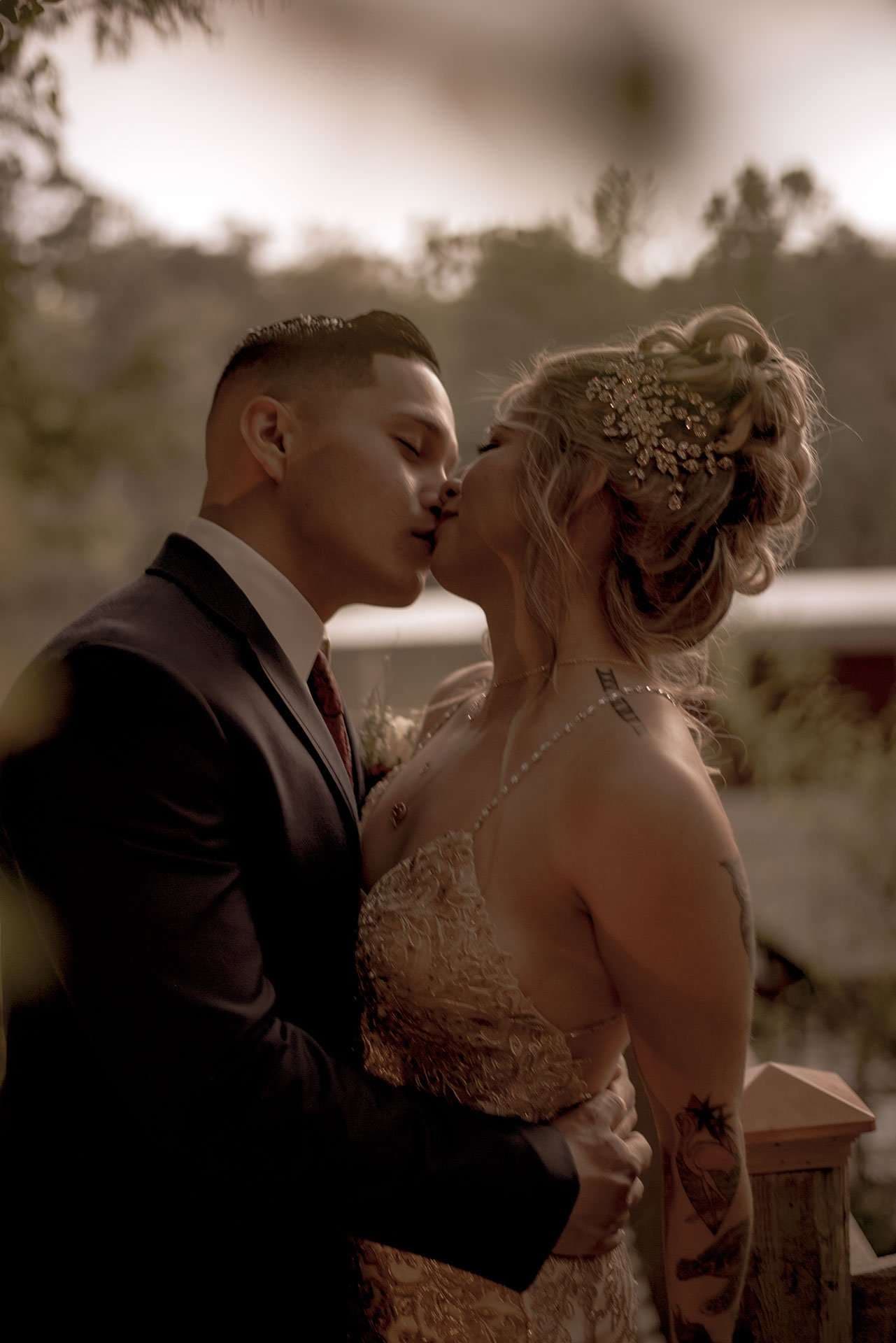 Sepia-toned couple kissing outdoors, man in dark suit and red tie, woman in gold gown with jeweled updo