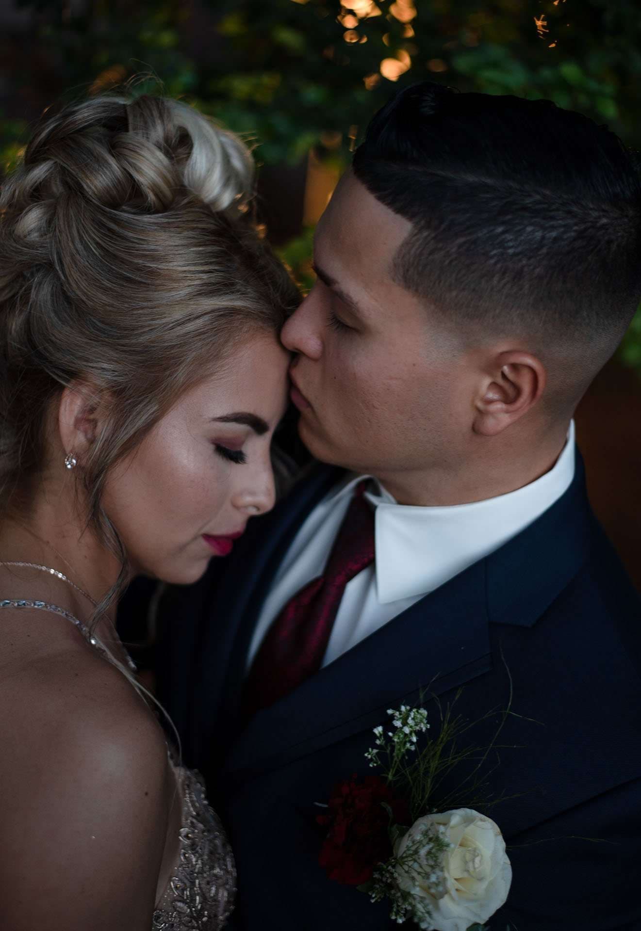 Man in a navy suit with a red tie and boutonniere kissing the forehead of a woman with an updo