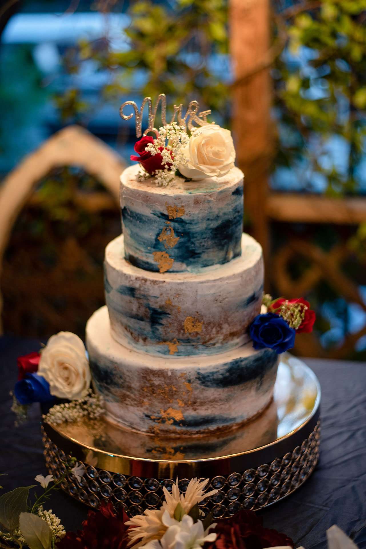 Three-tier wedding cake with blue and gold watercolor frosting, red and white roses, and a Mr & Mrs topper