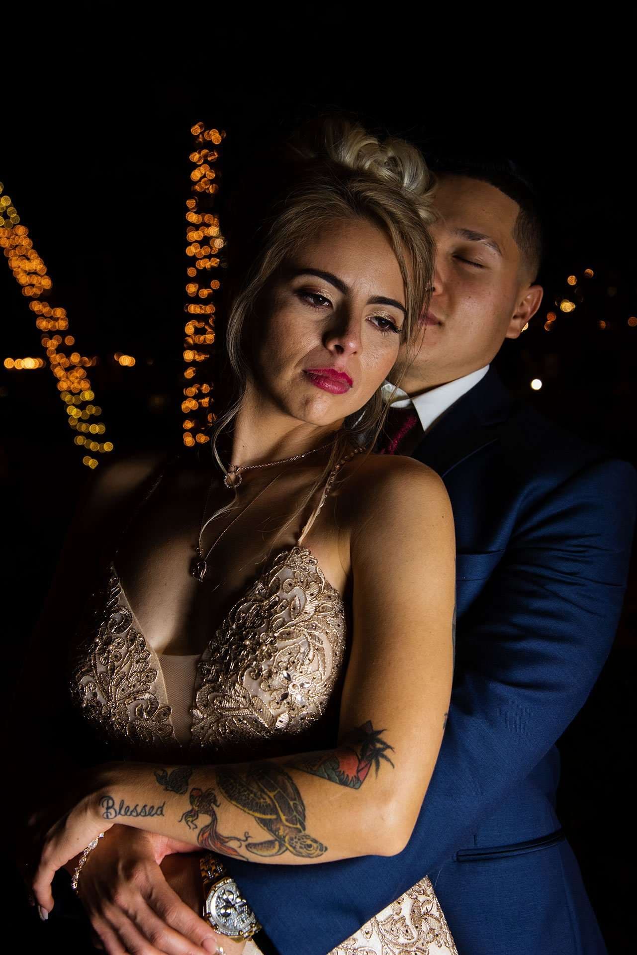 Groom in a blue suit embracing his bride from behind at night, string lights glowing behind them