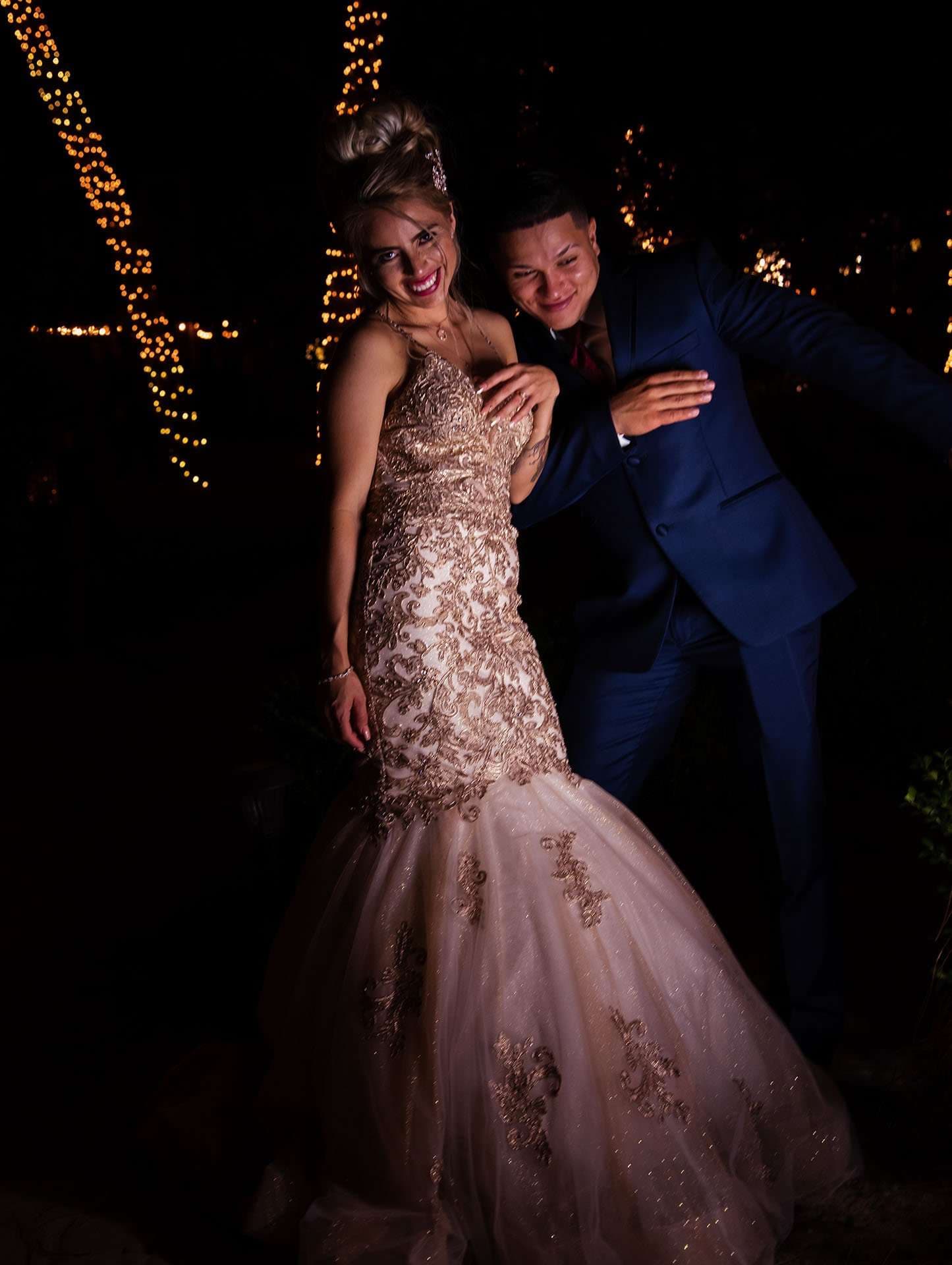 Couple laughing at night beside string lights, woman in a gold lace mermaid gown, man in a blue suit