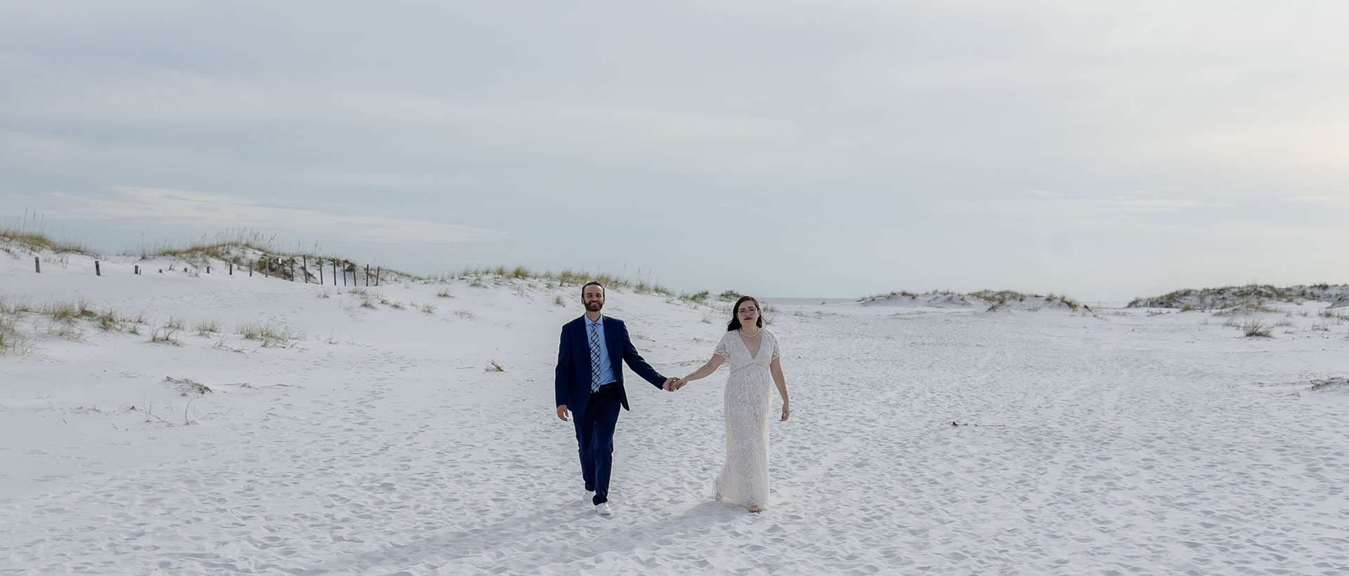 Couple in a navy suit and white lace gown holding hands while walking across white sand dunes on Okaloosa Island