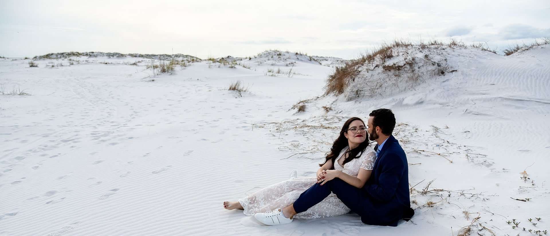 Couple in wedding attire sitting close together on white sand dunes at Okaloosa Island, bride barefoot in a lace dress