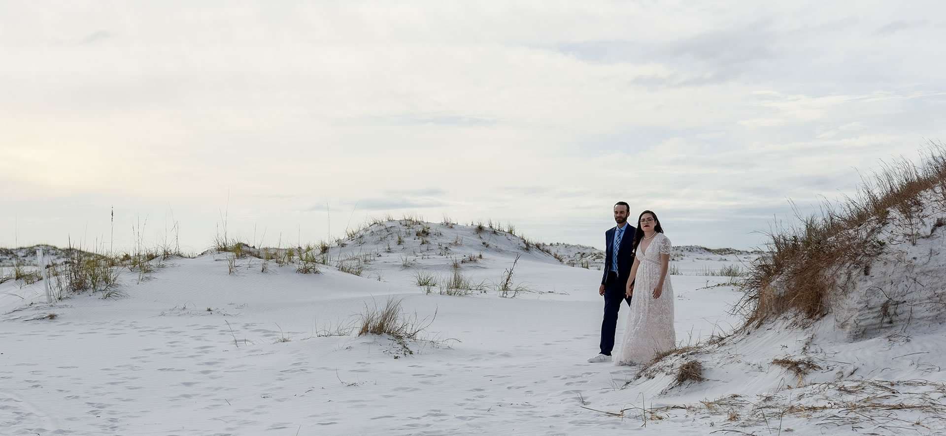 Couple in a navy suit and white lace gown standing among white sand dunes on Okaloosa Island at dusk
