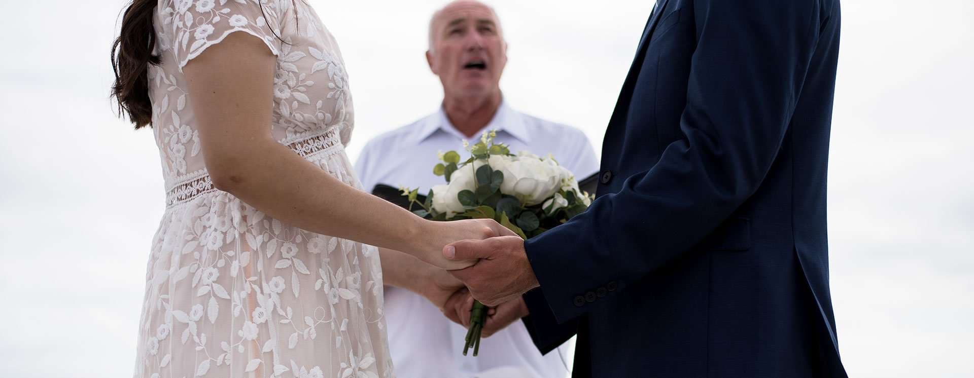 Couple holding hands during their ceremony on Okaloosa Island, white rose bouquet between them and officiant behind
