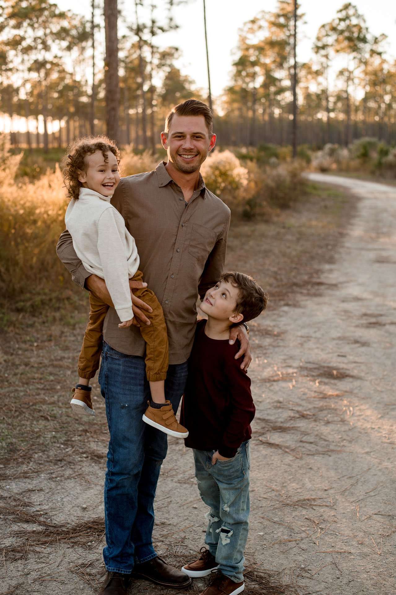 Smiling man holding one laughing young boy while another boy looks up at him on a sandy path at sunset