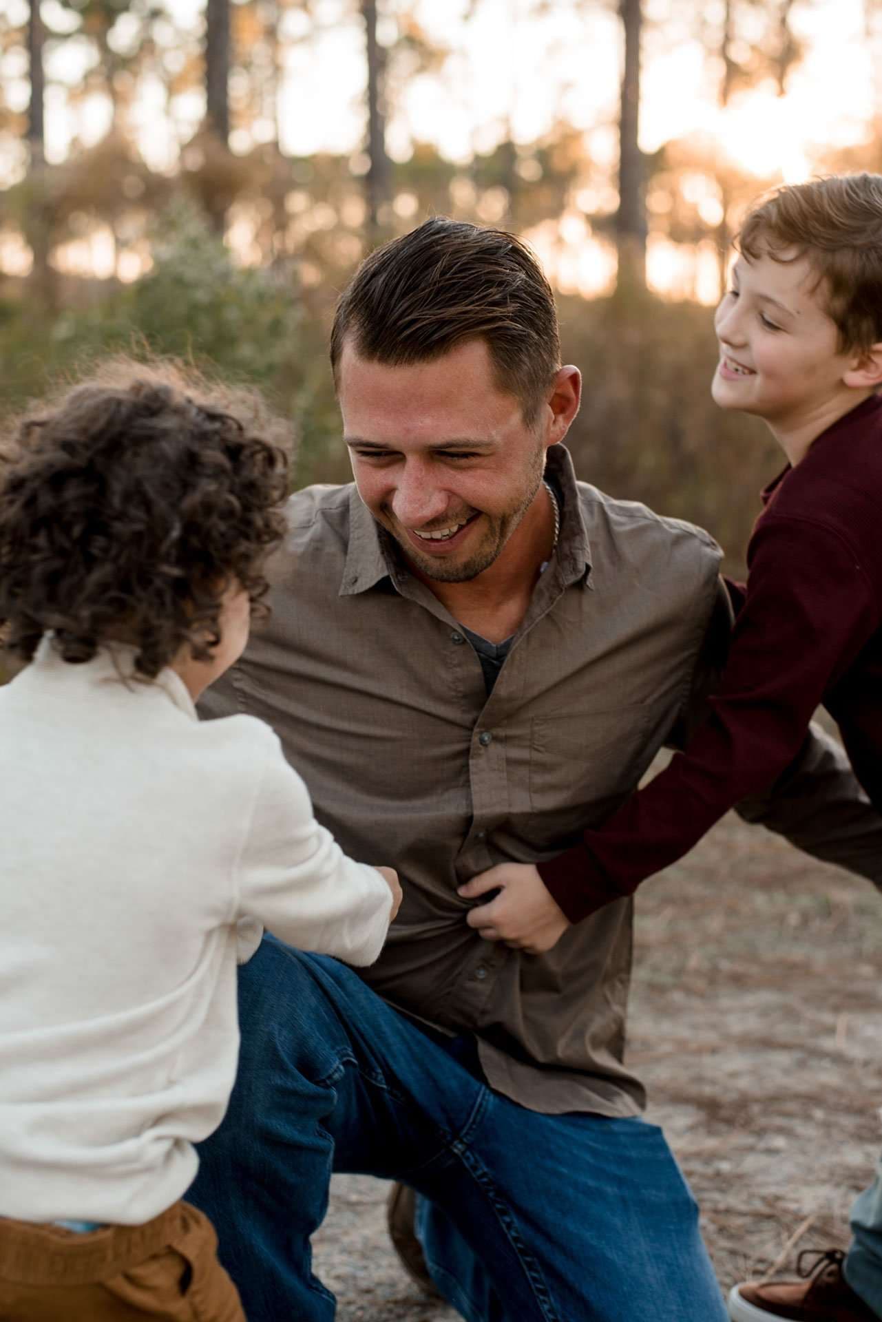Smiling man crouching outdoors as two laughing boys playfully lean in around him at sunset in the woods