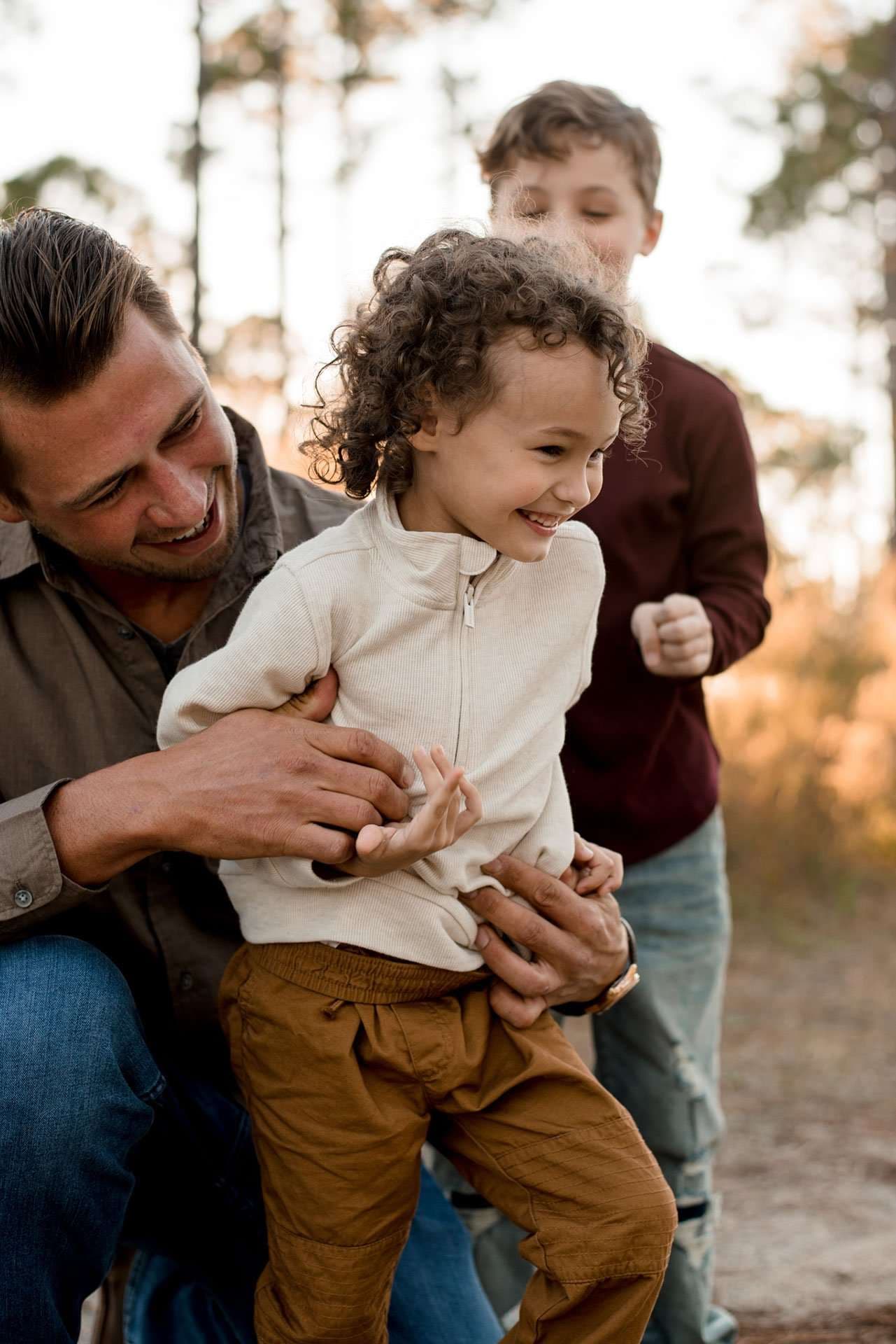 Smiling man holding a laughing curly-haired child while an older boy stands behind them in warm autumn light