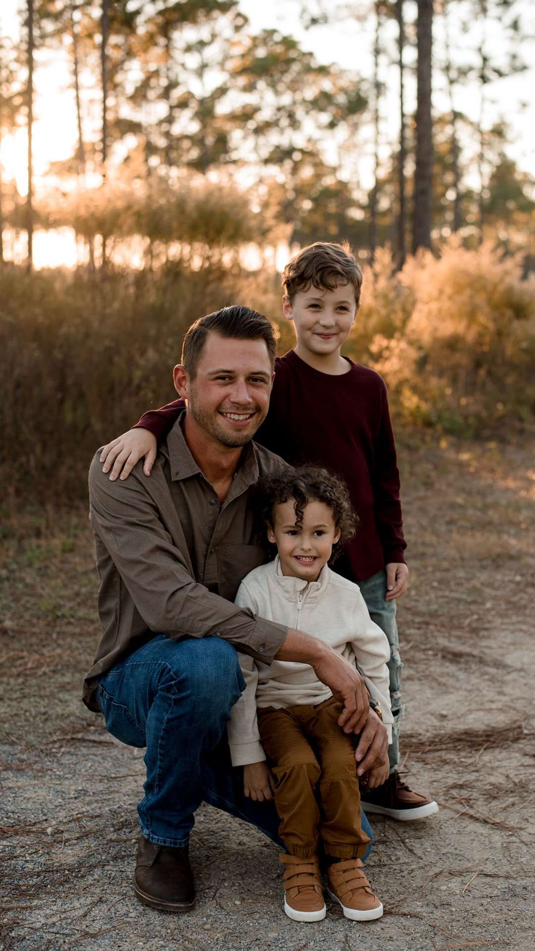 Smiling man crouching with two young boys in front of him on a sandy path, warm sunset light through pine trees