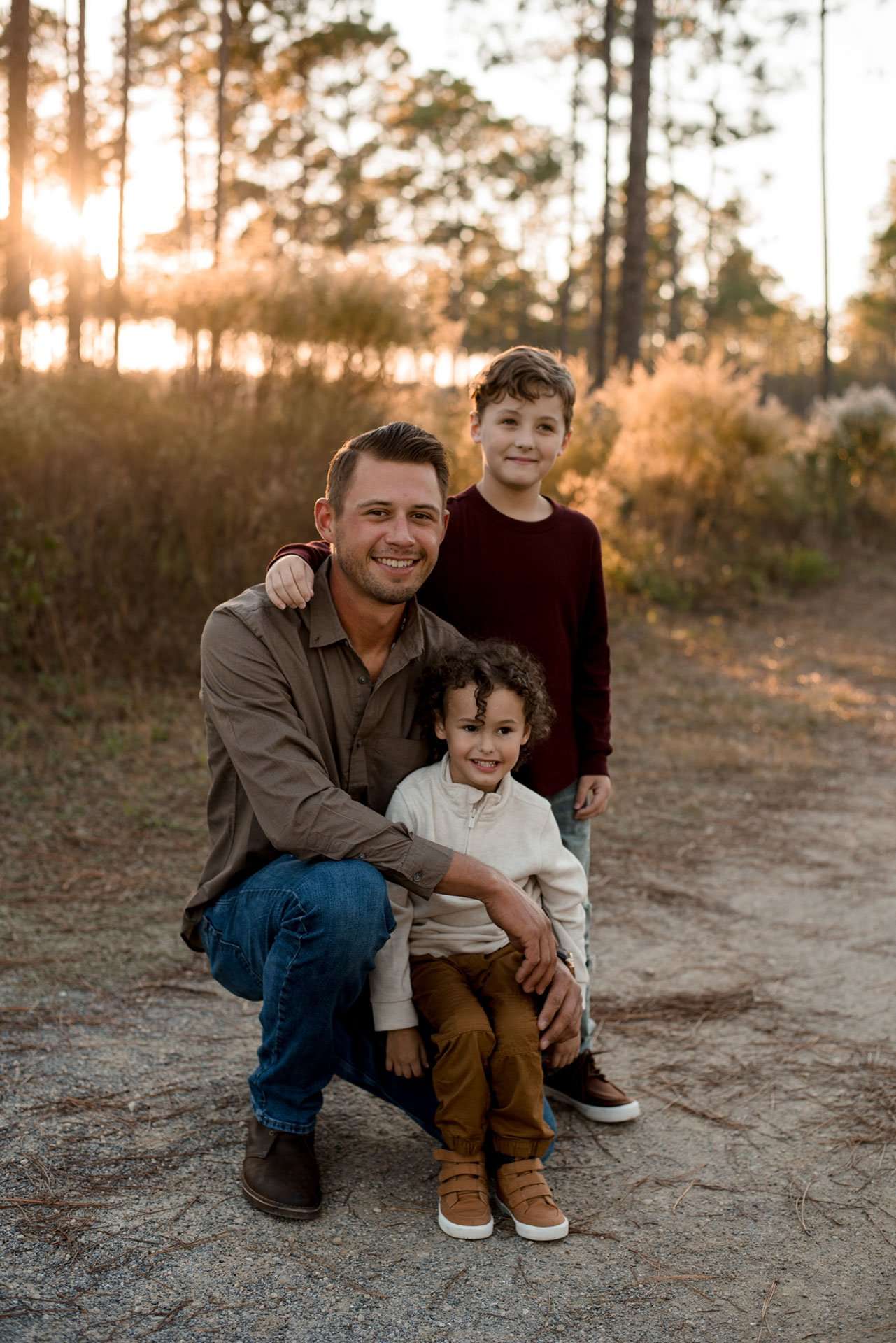 Smiling man crouching with two young boys on a sandy path in golden sunset light among pine trees