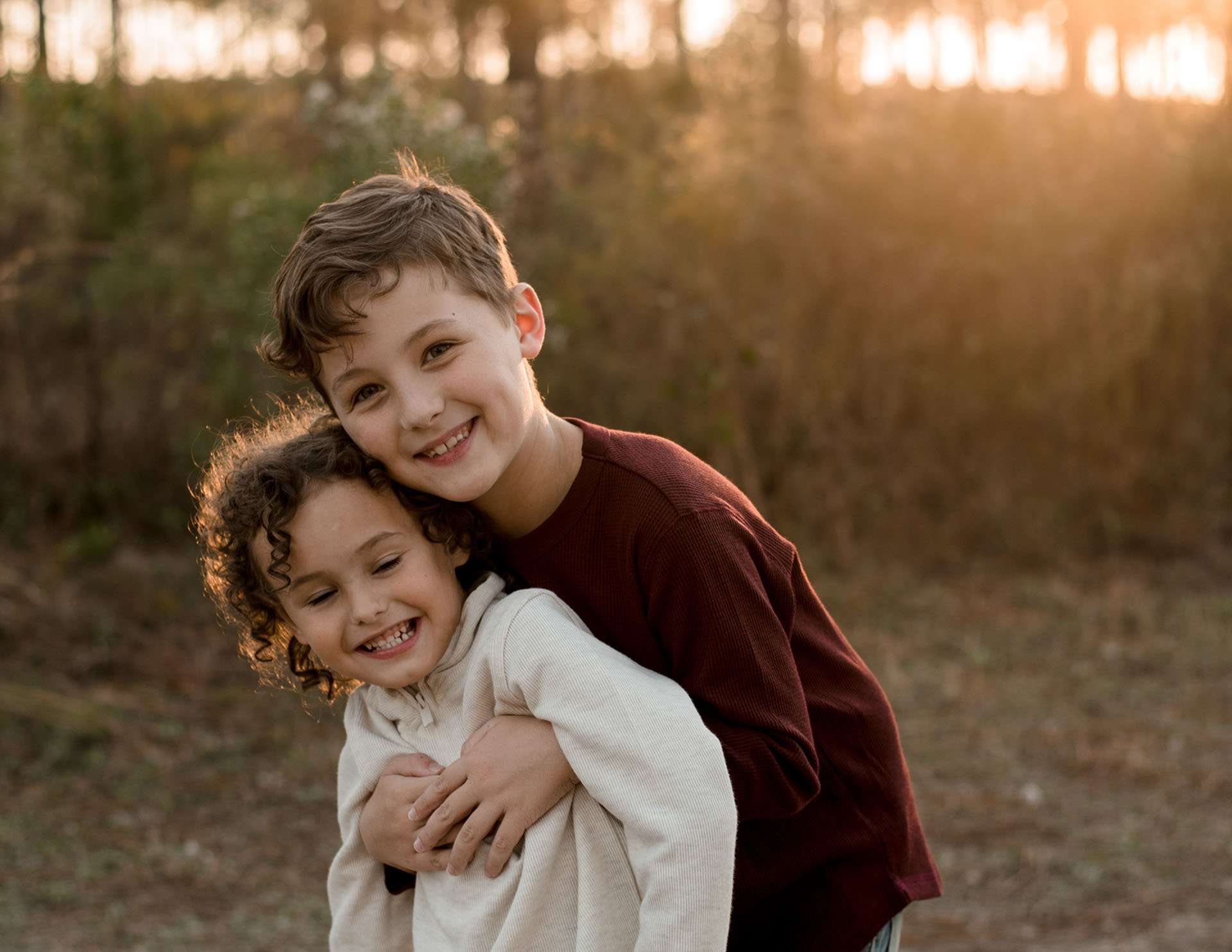 Two smiling boys hugging outdoors, one in a maroon shirt embracing a curly-haired boy from behind in golden light