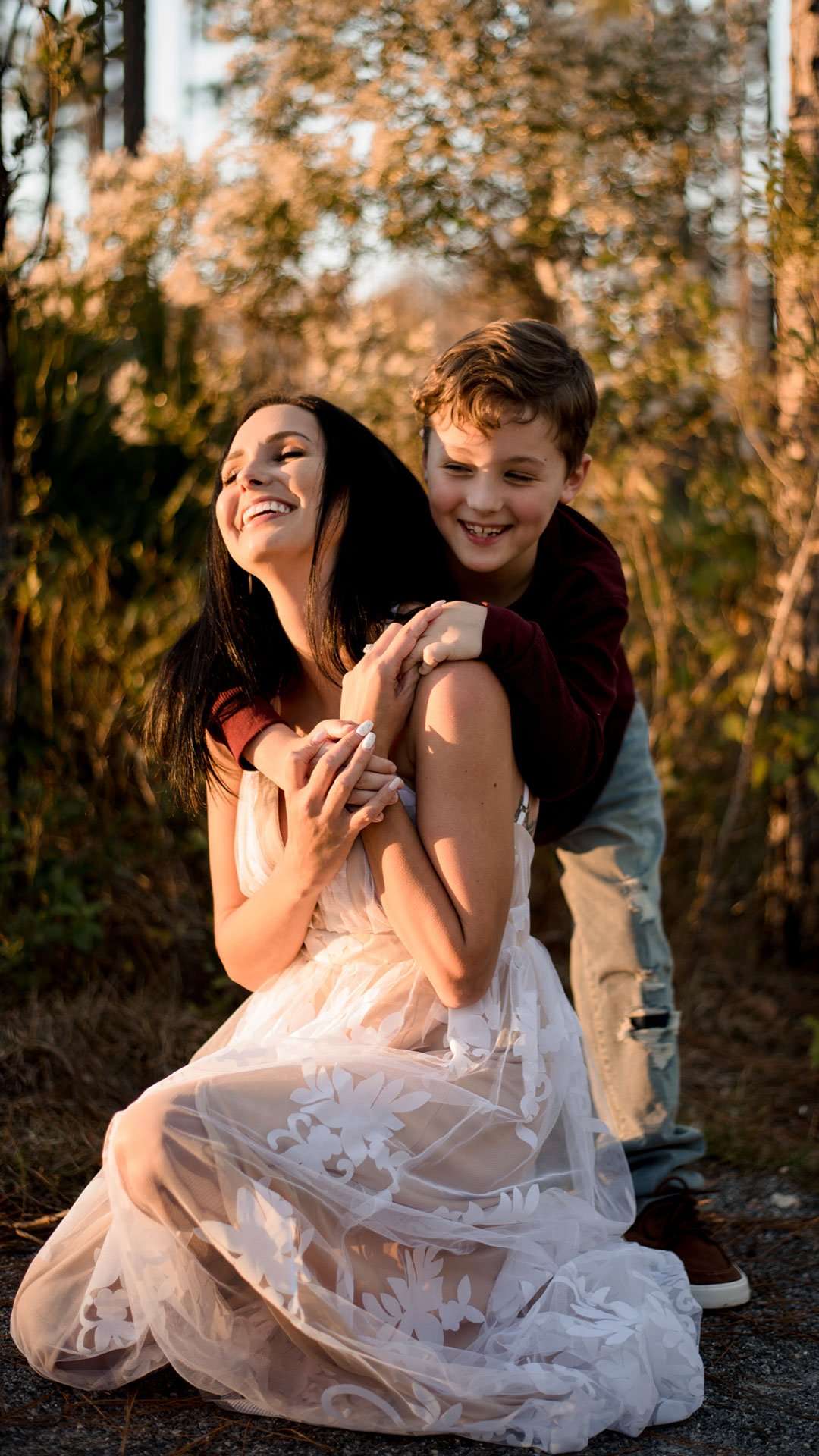 Smiling boy hugging a laughing woman in a white lace dress from behind among golden autumn grasses