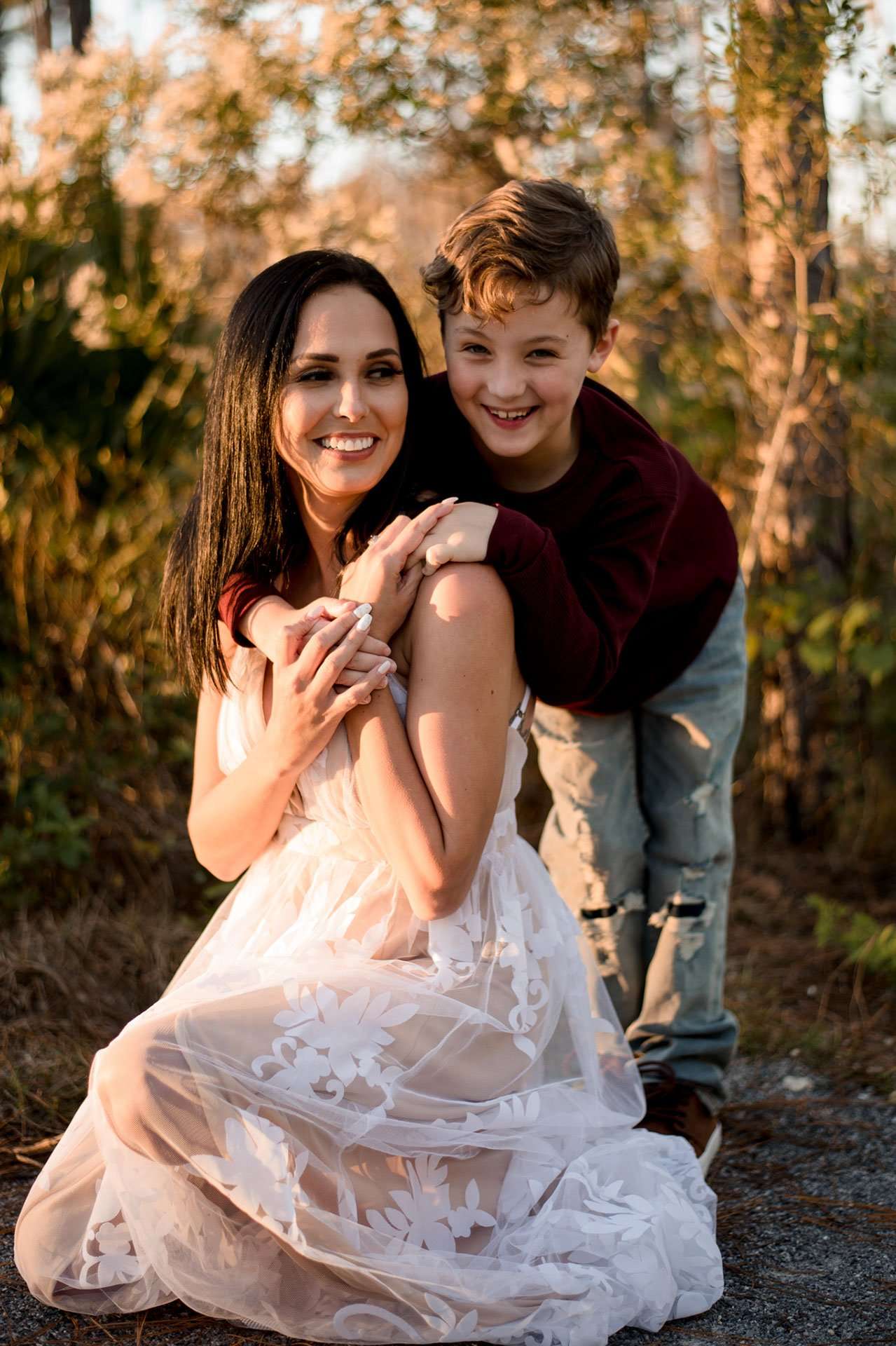 Smiling woman in a white lace dress with a laughing boy hugging her from behind among golden autumn foliage