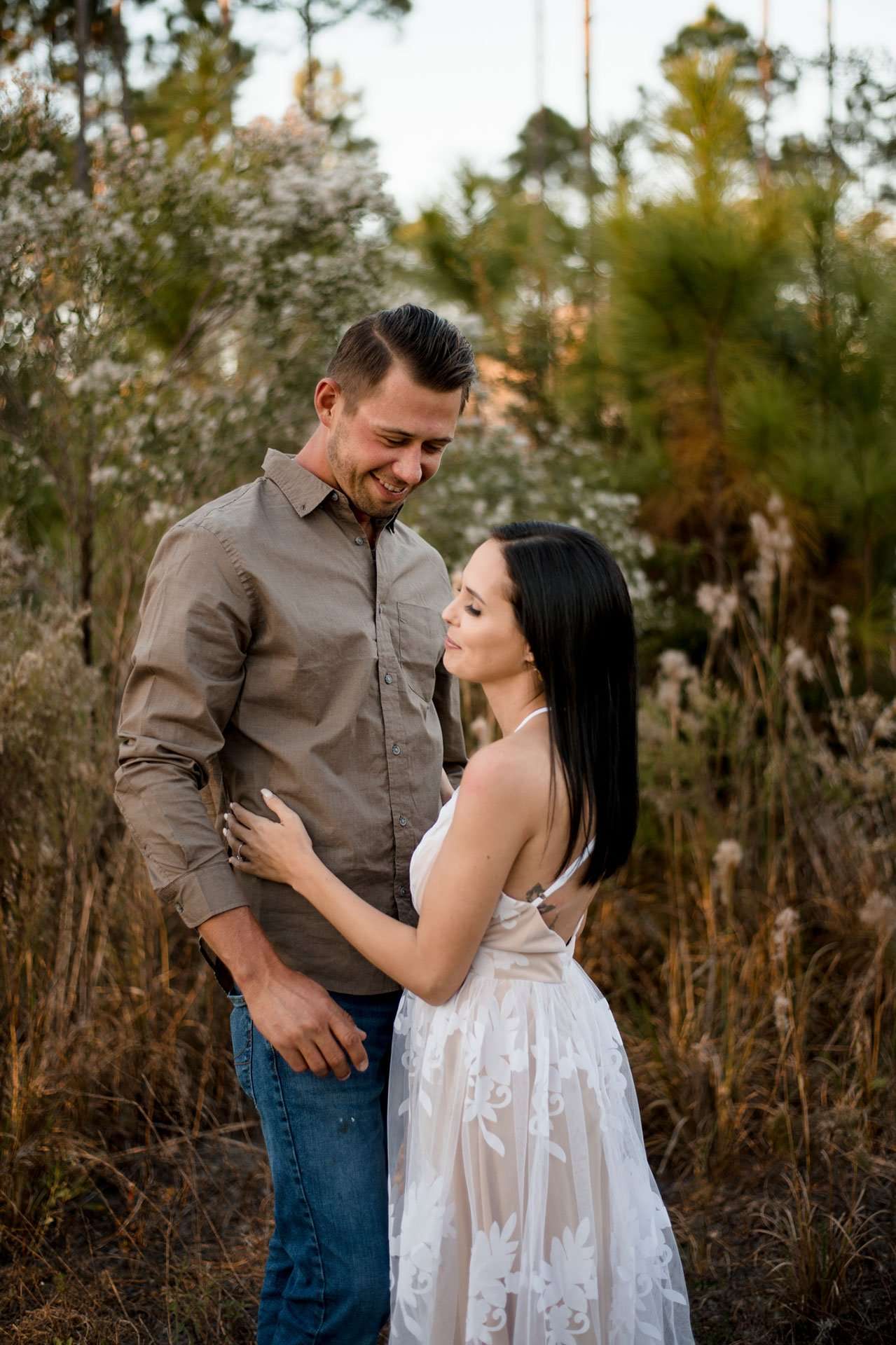 Smiling couple embracing in a grassy field of autumn wildflowers, woman in a white lace dress, man in an olive shirt