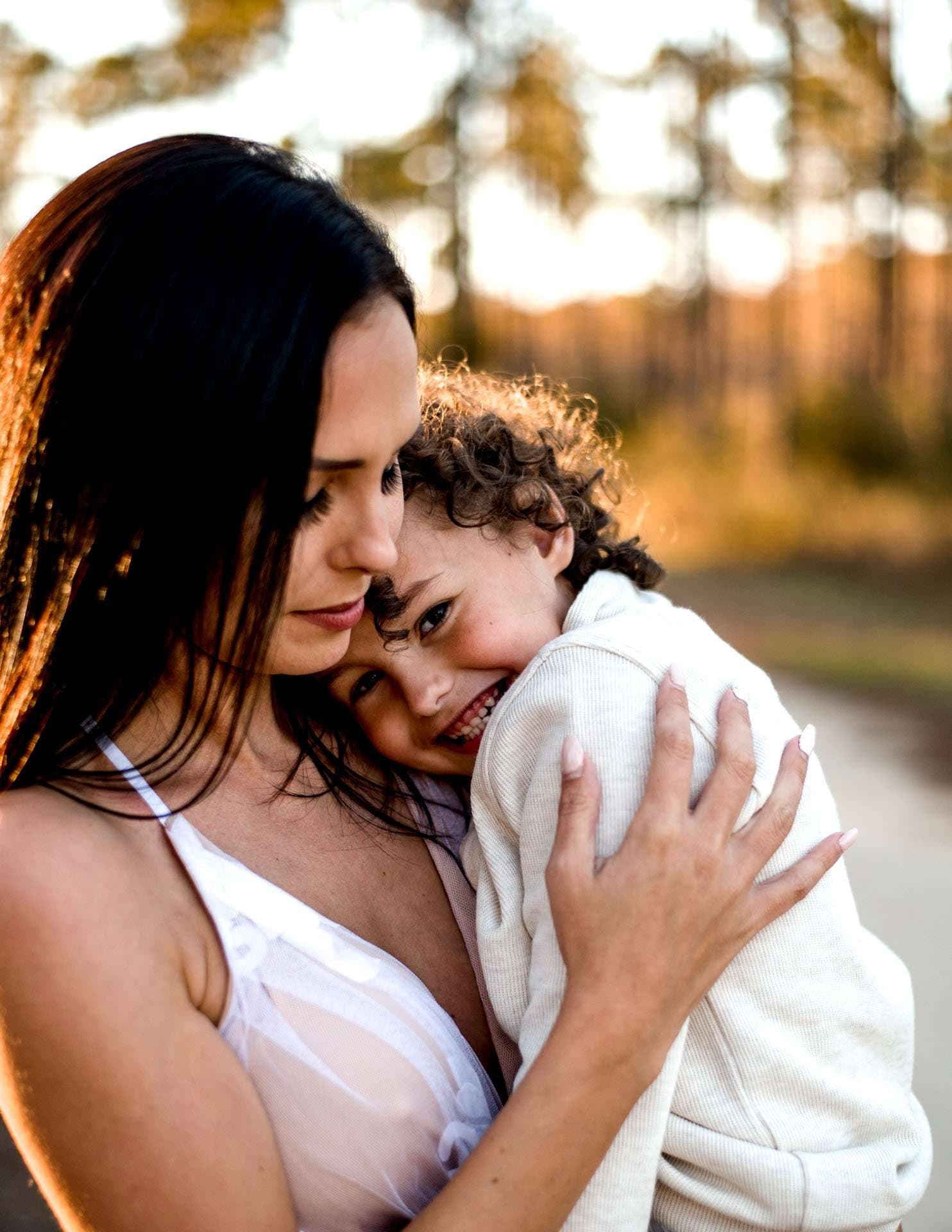 Smiling curly-haired child hugged close by a dark-haired woman in white, warm golden light on a wooded path