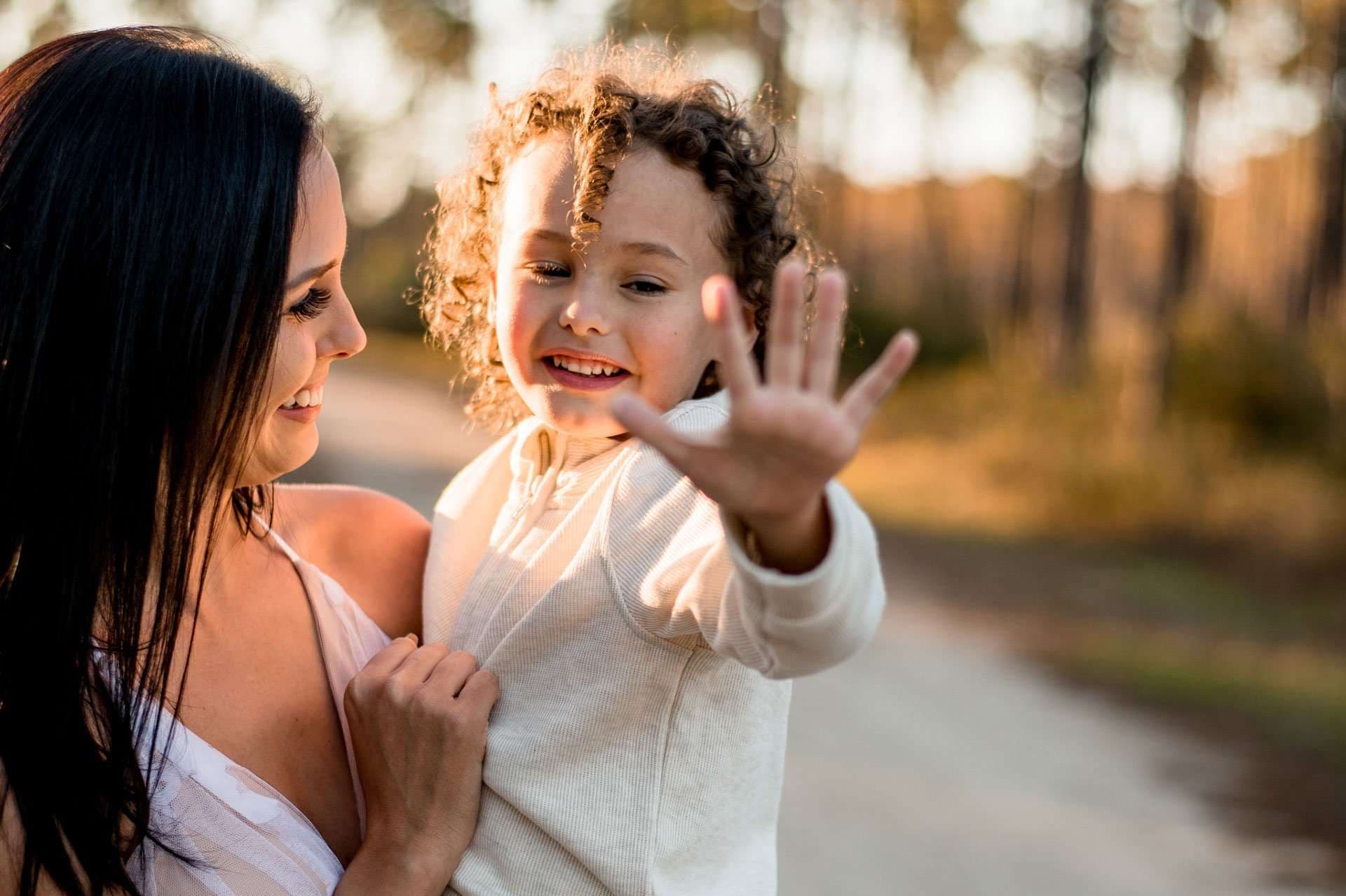 Smiling woman holding a curly-haired child who waves an open hand toward the camera on a sunlit forest path