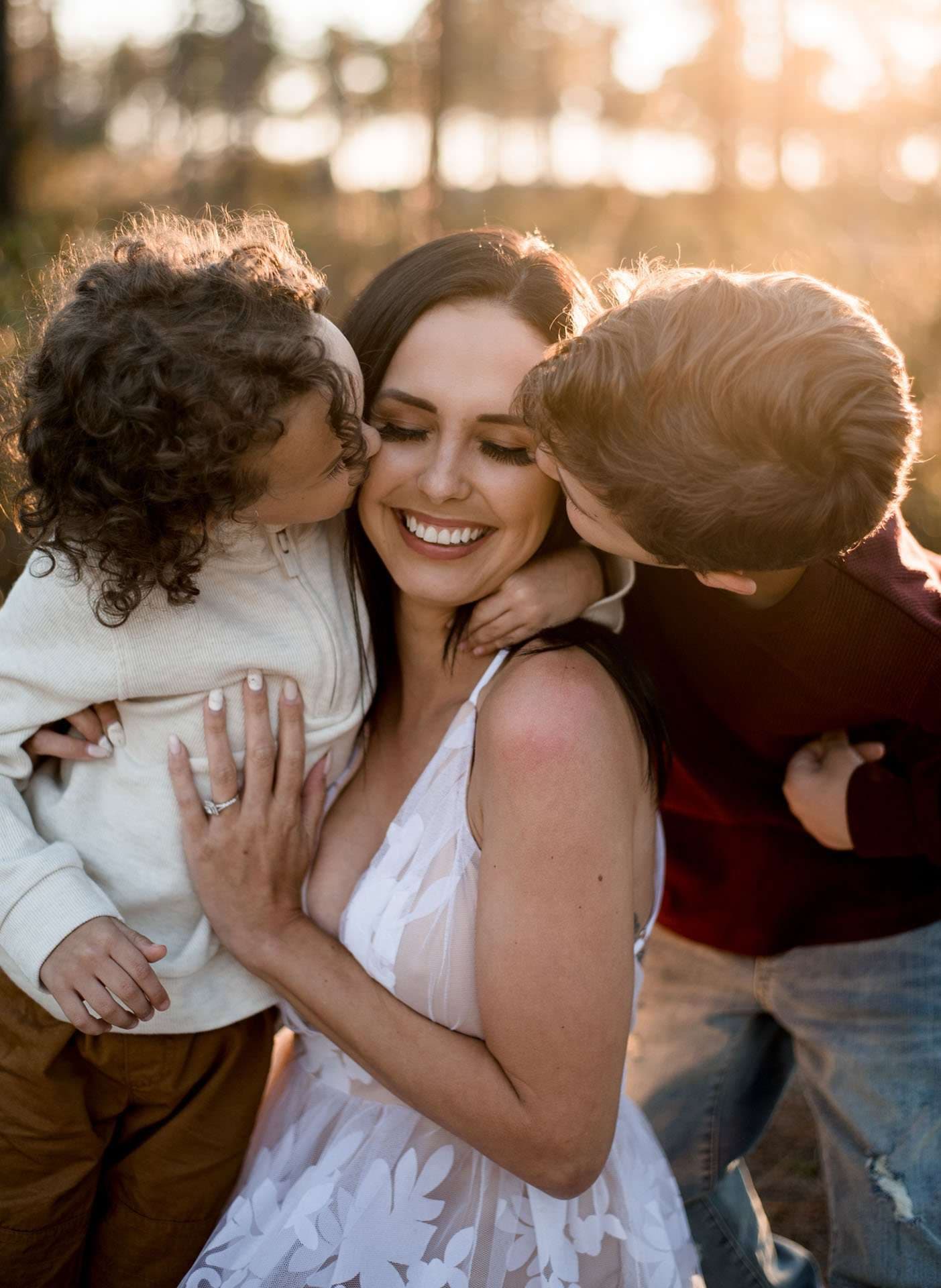 Smiling woman in a white lace dress is kissed on both cheeks by two children in golden backlight outdoors