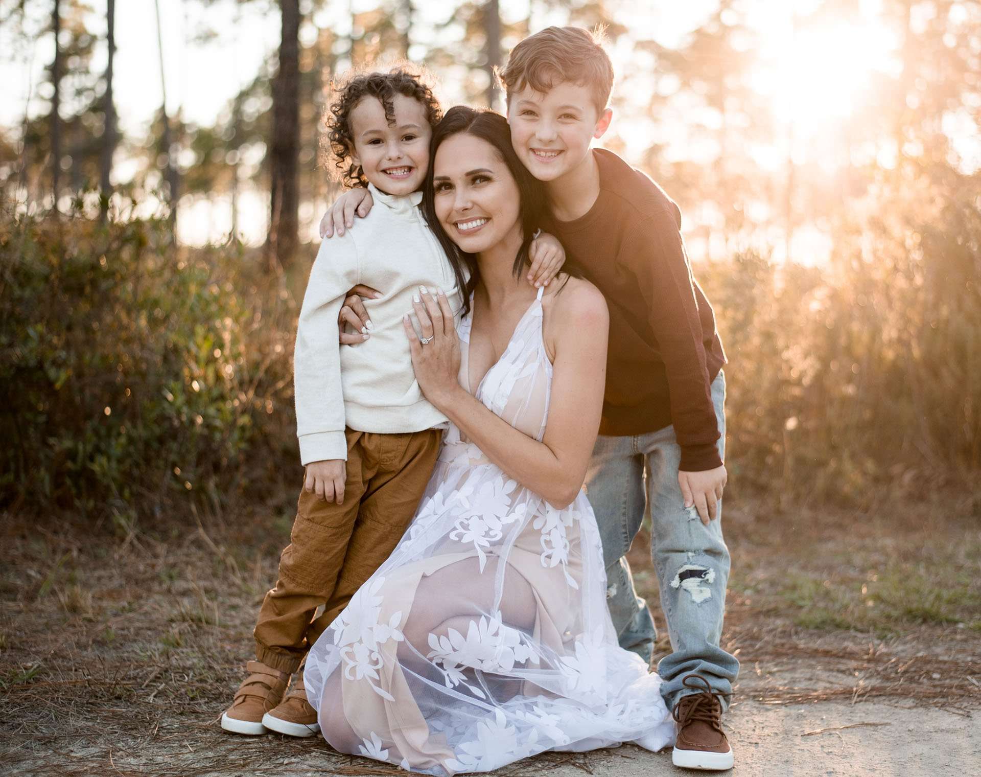 Smiling woman in a white lace gown embraced by two boys in a sunlit pine forest at Conservation Park