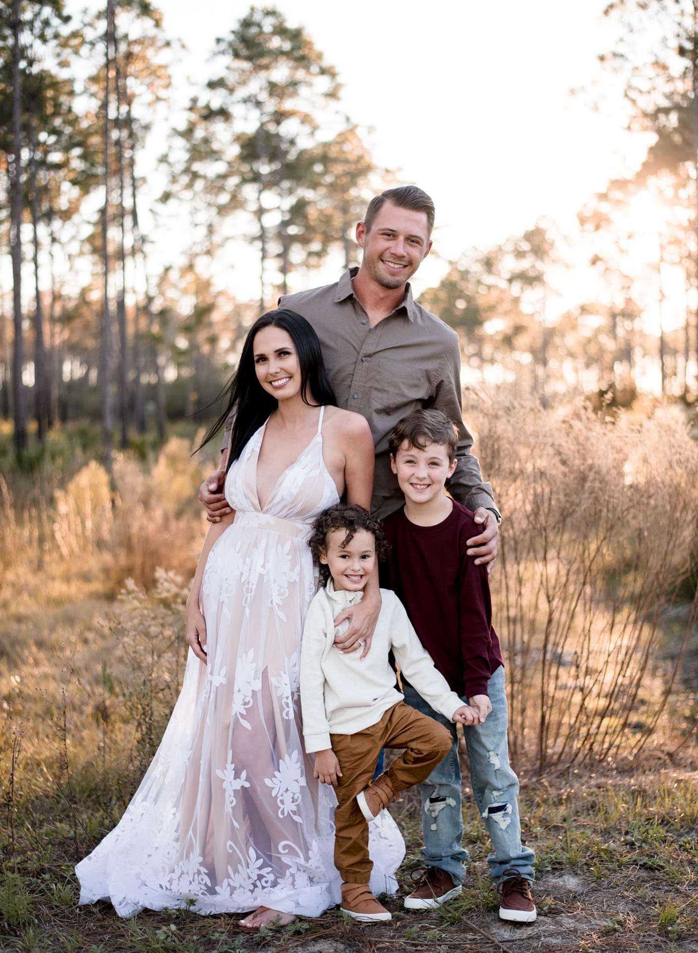 Smiling family of four posing close together in a golden autumn pine forest, mother in a white lace gown