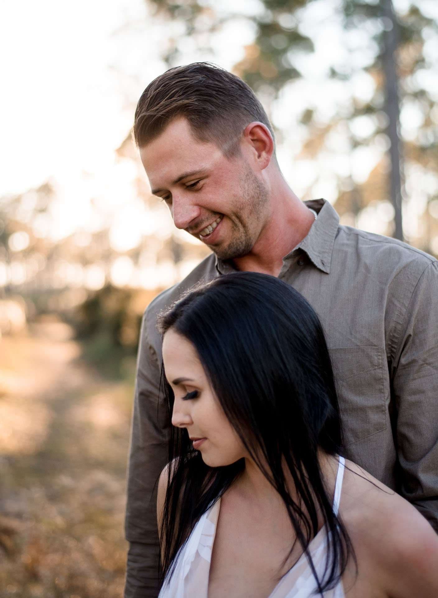 Smiling man in an olive button-up shirt looks down at a dark-haired woman in white in golden-lit woods