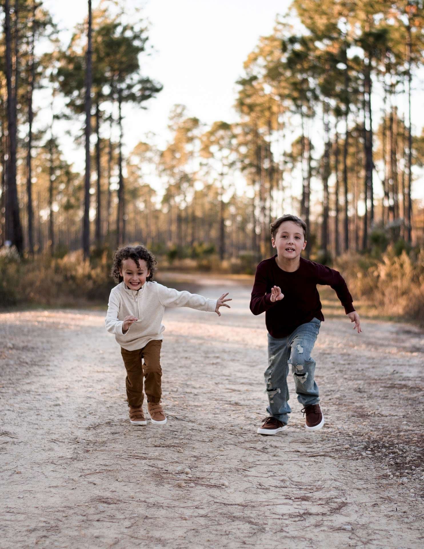 Two smiling young boys running along a sandy path through tall pine trees at Panama City Beach Conservation Park
