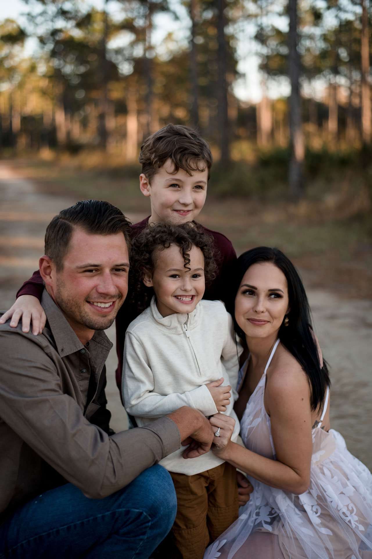 Family of four smiling close together on a sandy trail among tall pines in autumn light