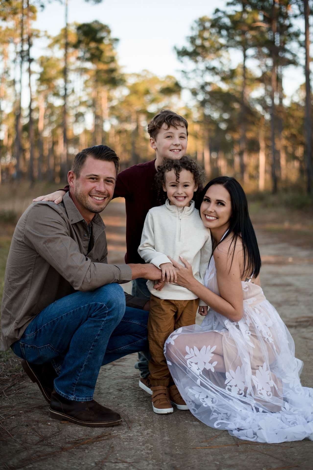 Smiling family of four huddled together on a sandy forest trail, woman in a white lace gown, tall pines behind