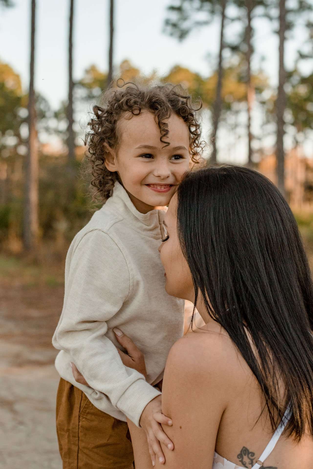 Smiling curly-haired child in a cream sweater hugged by a dark-haired woman among tall pine trees at the conservation park