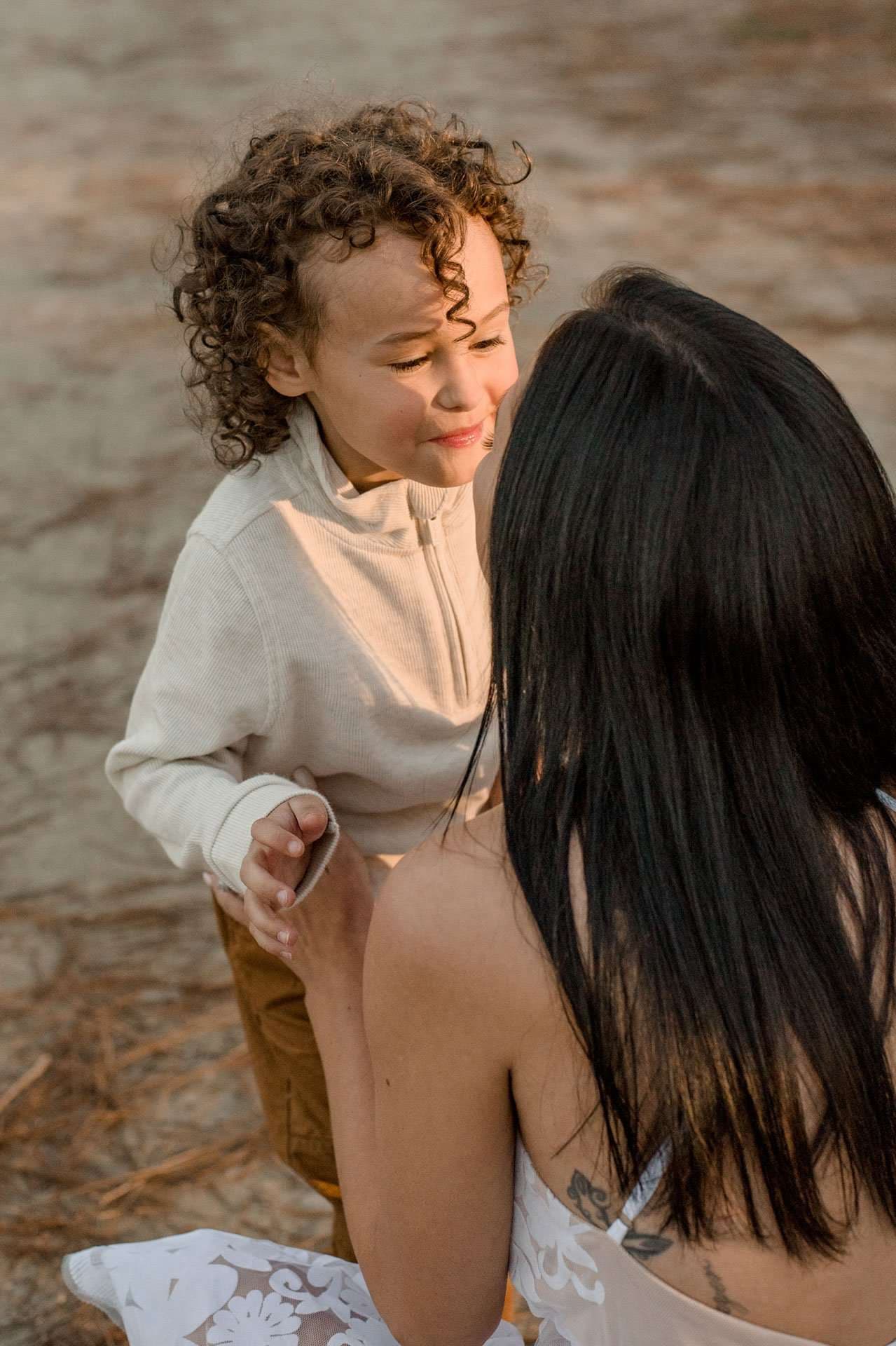 Smiling curly-haired child in a cream zip pullover leans toward a long-haired woman in a white lace top at Conservation Park