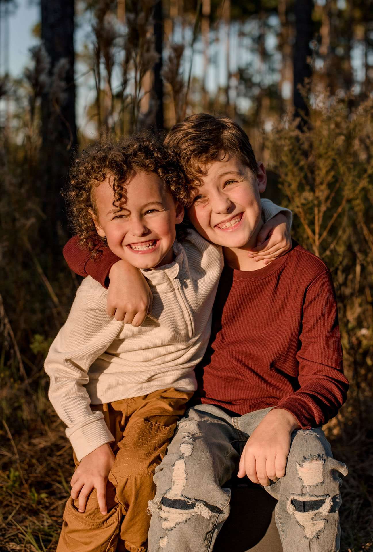 Two smiling boys hugging in warm golden light, one with curly hair in cream and one in a rust sweater, in the woods