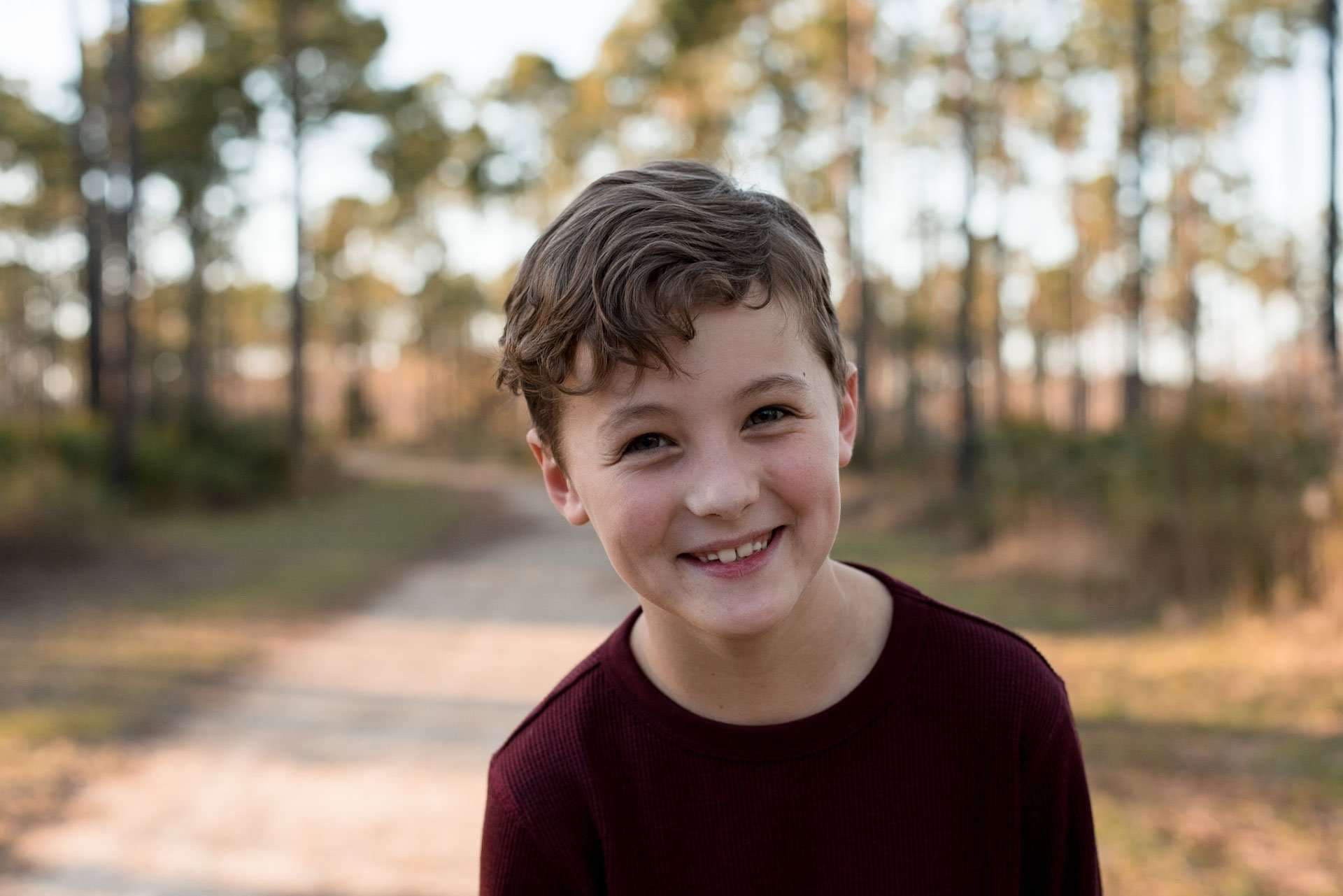 Smiling boy in a maroon sweater standing on a sandy trail with pine trees softly blurred behind him