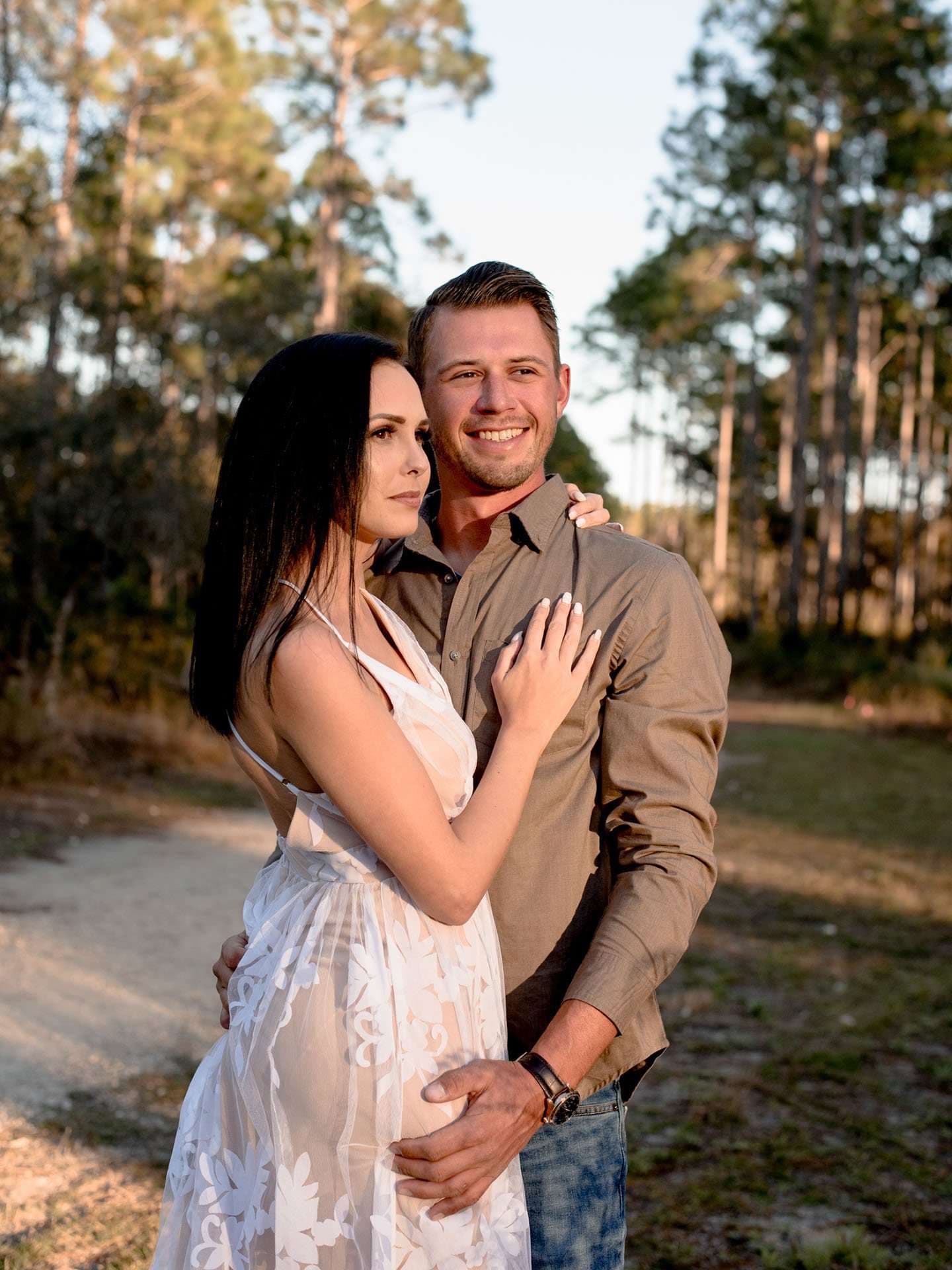 Couple embracing on a sandy trail among tall pines, woman in a white lace sundress
