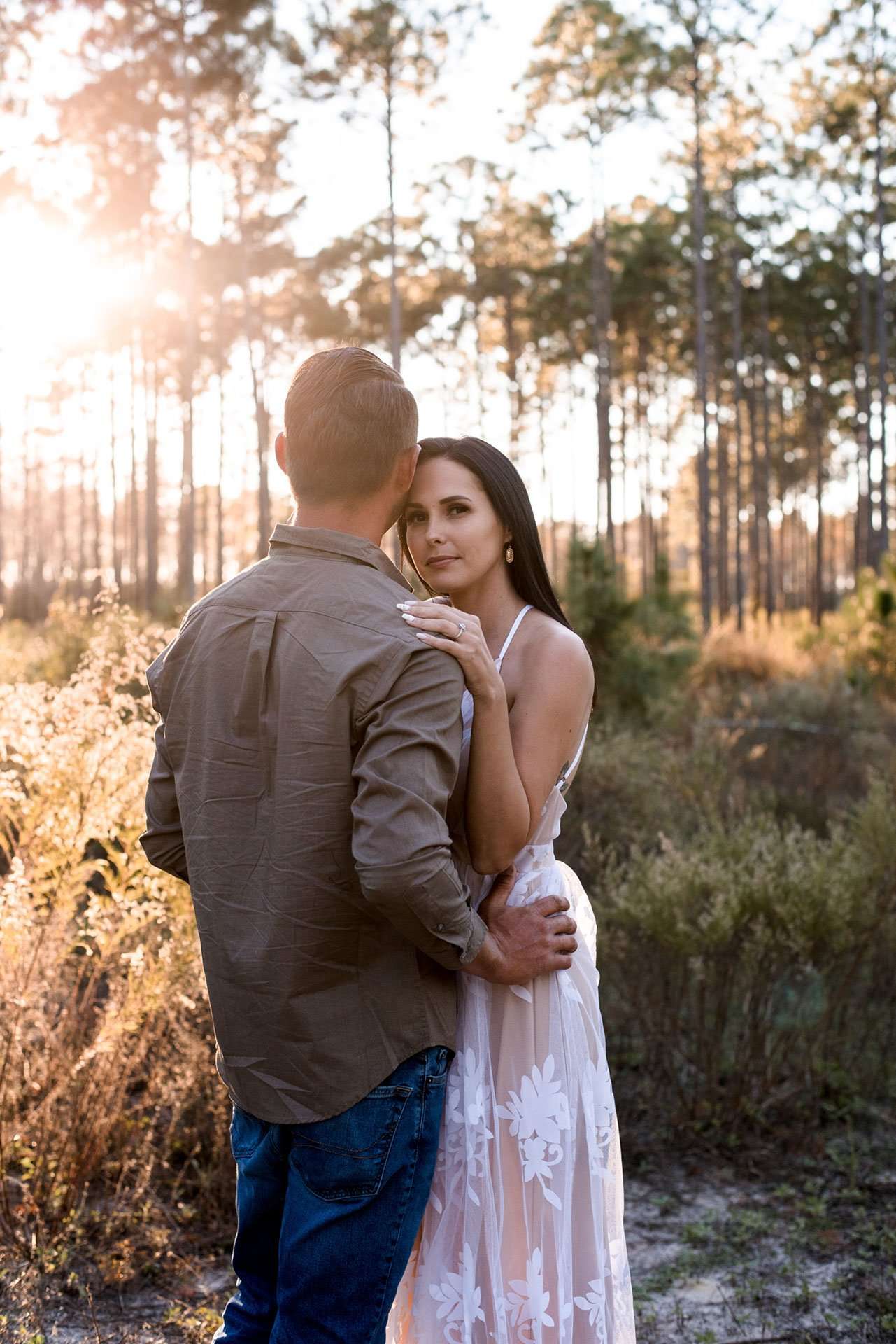 Couple embracing in a sunlit pine forest at golden hour, woman in a white lace dress resting her hand on his shoulder