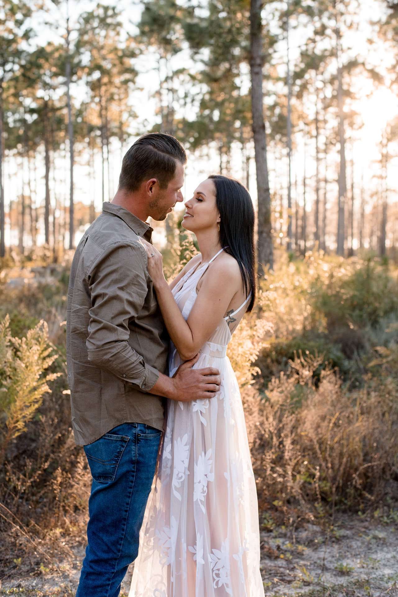 Couple embracing and almost kissing in a sunlit pine forest, woman in a lace-detailed white dress, man in olive shirt