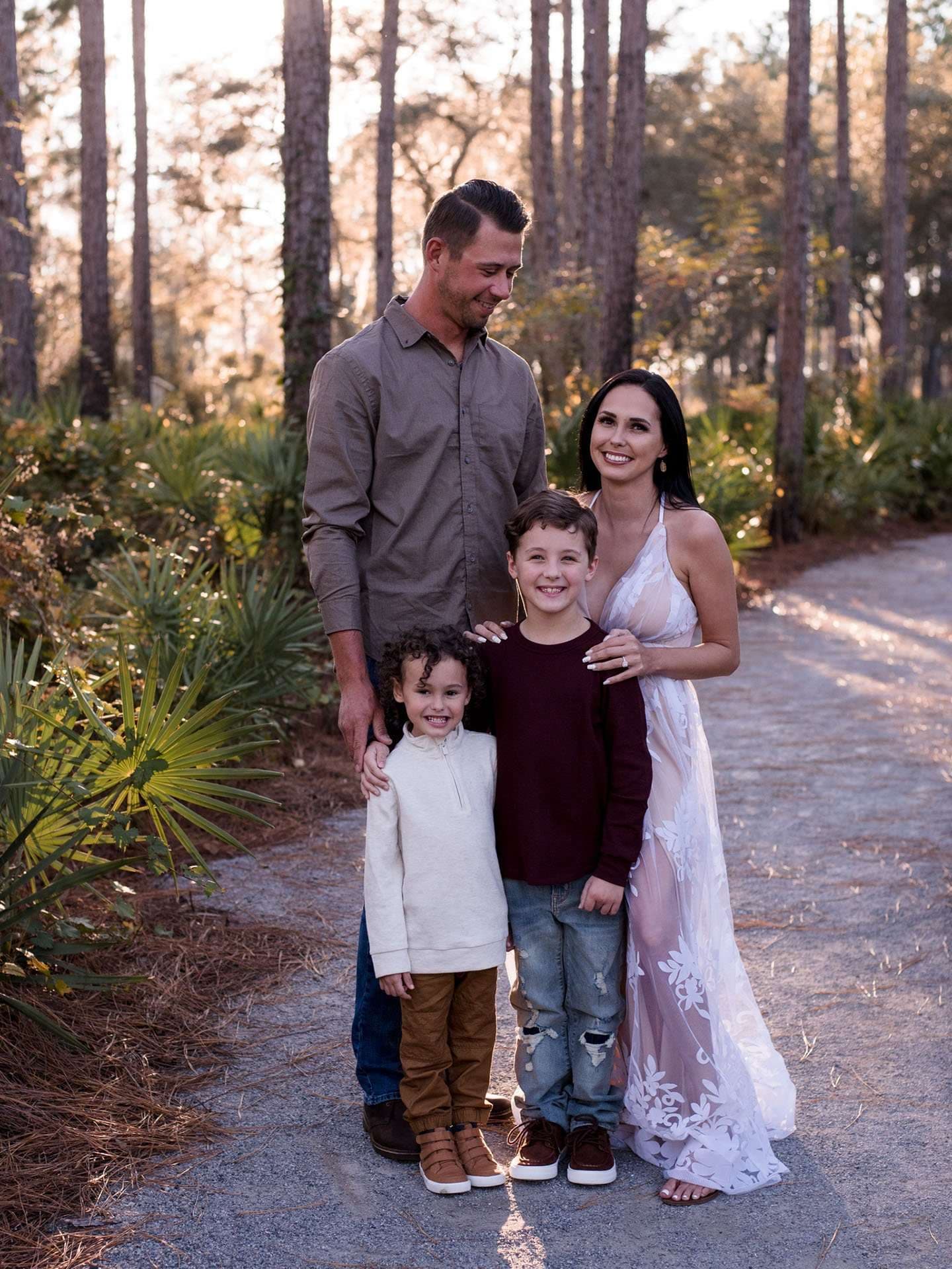Smiling family of four, parents and two young boys, posed together on a sandy path among pine trees at sunset