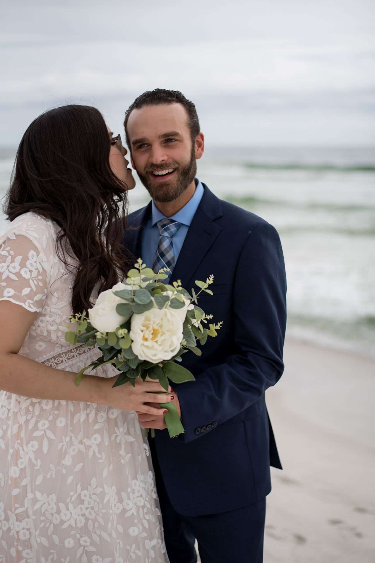 Smiling groom in a navy suit holding a white rose bouquet as his bride kisses his cheek on Okaloosa Island beach