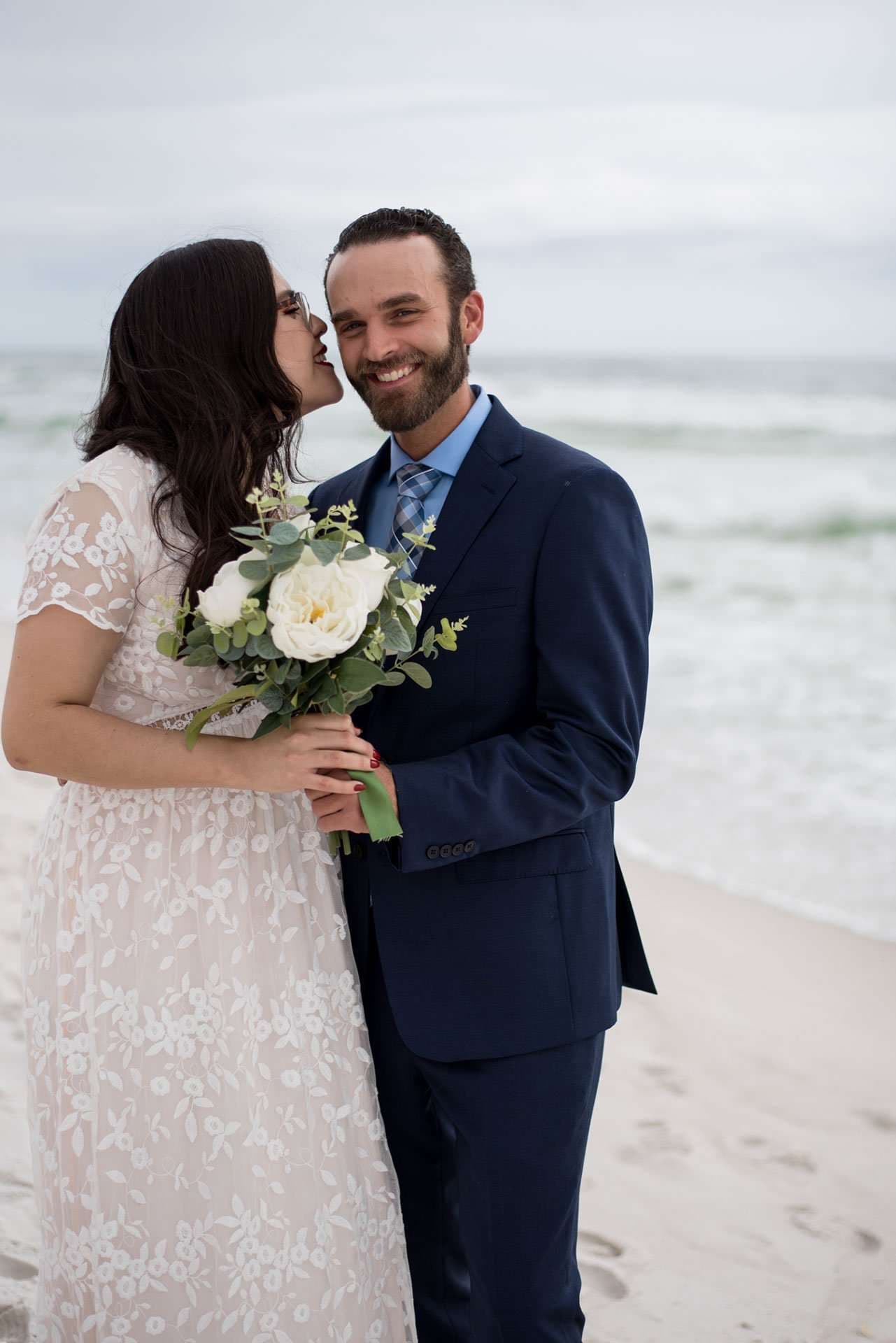 Smiling groom in navy suit holding a white rose bouquet as his bride whispers in his ear on the beach