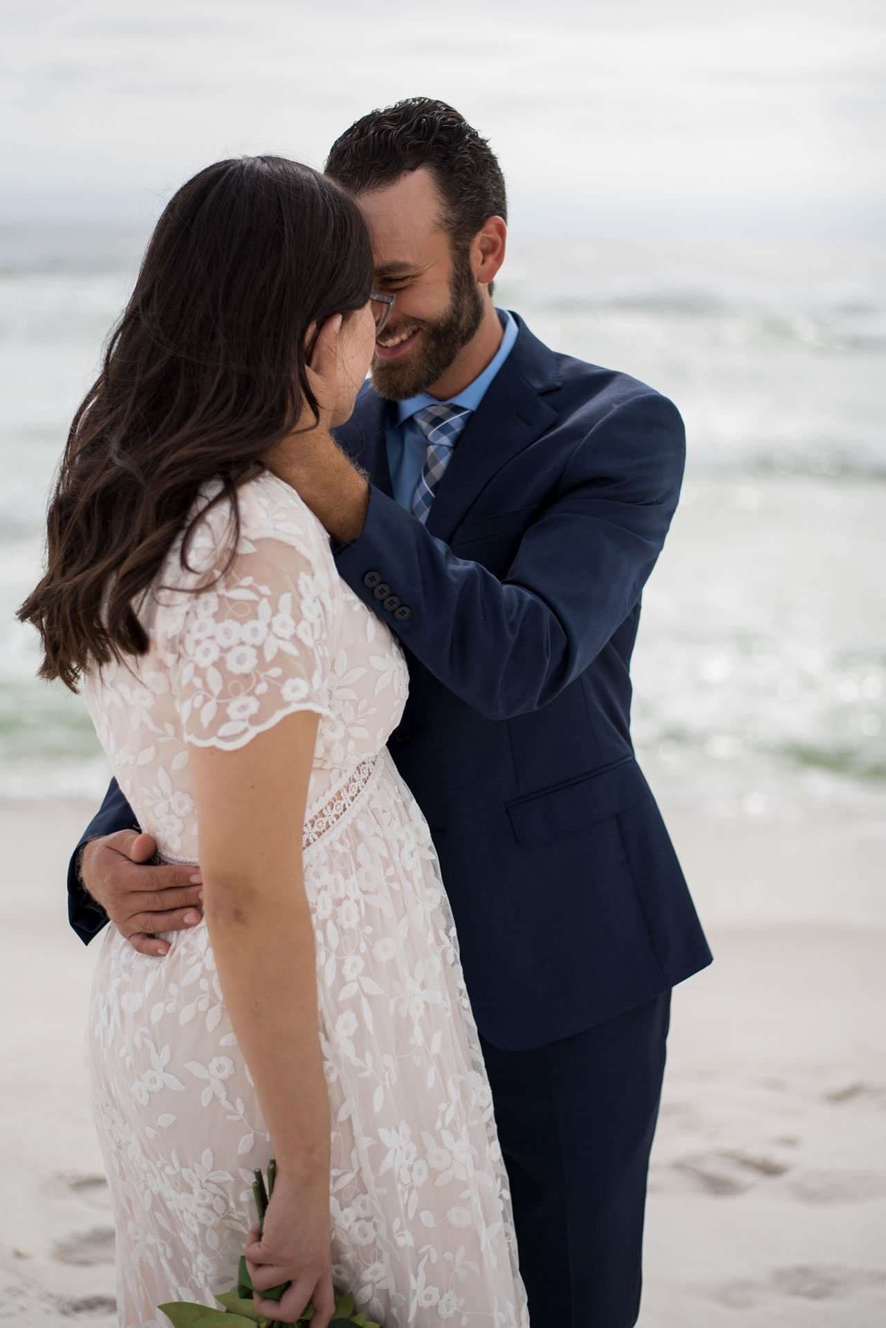 Smiling groom in a navy suit embraces his bride in a white lace dress on the sandy Okaloosa Island beach