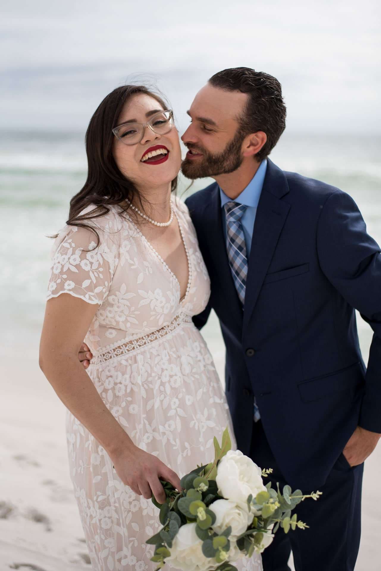 Laughing bride in a lace dress holding a white bouquet as her groom kisses her cheek on the beach