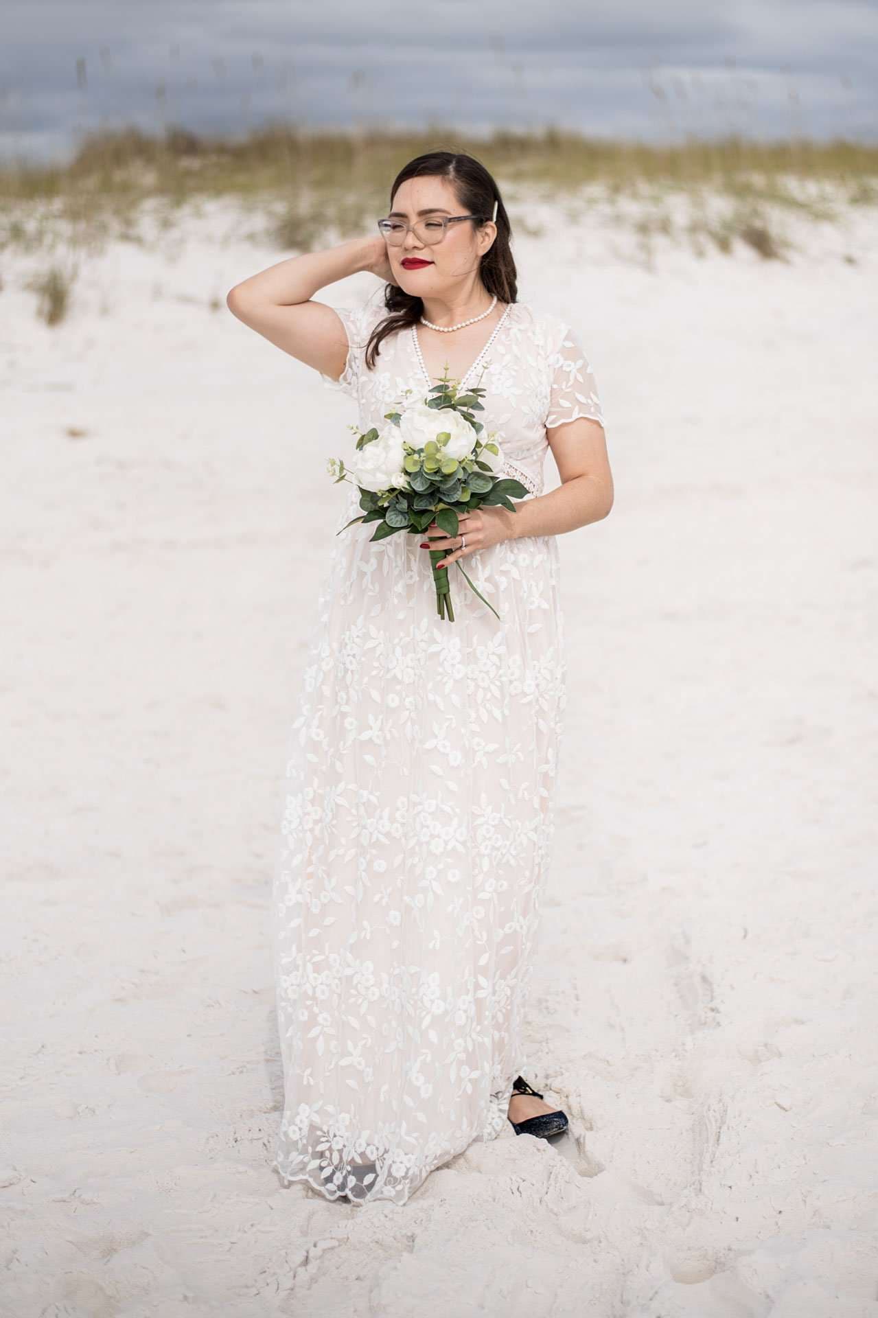 Bride in a white lace gown holding a white and green bouquet on Okaloosa Island's sandy dunes