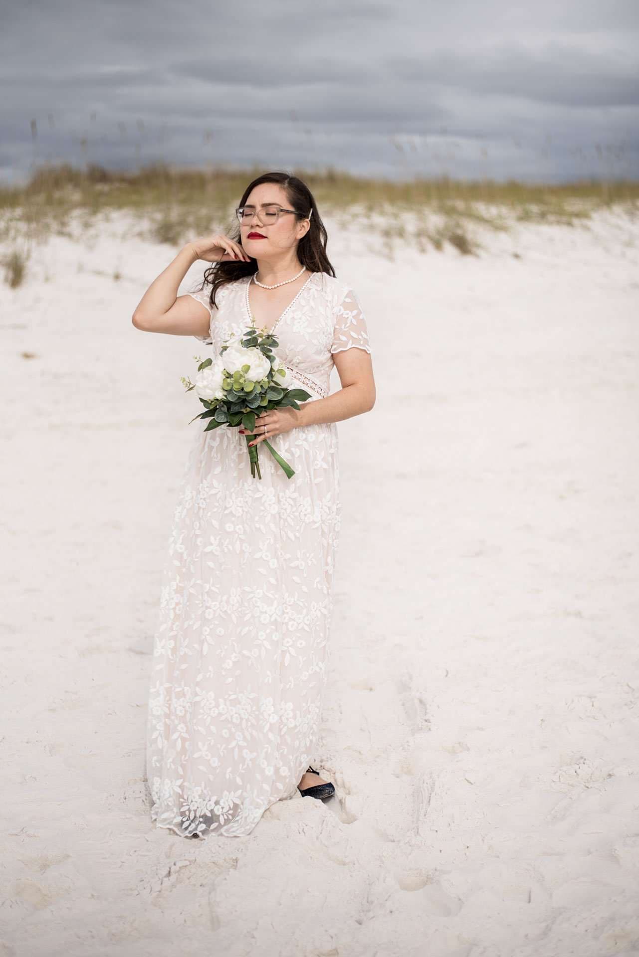 Bride in a white lace gown holding a white bouquet on the sand dunes under a stormy sky