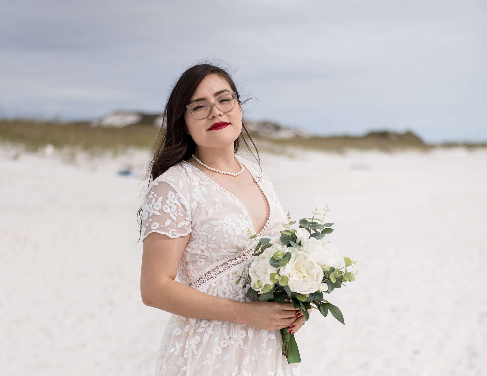 Bride in a white lace dress and glasses holding a white rose bouquet on the white sand at Okaloosa Island